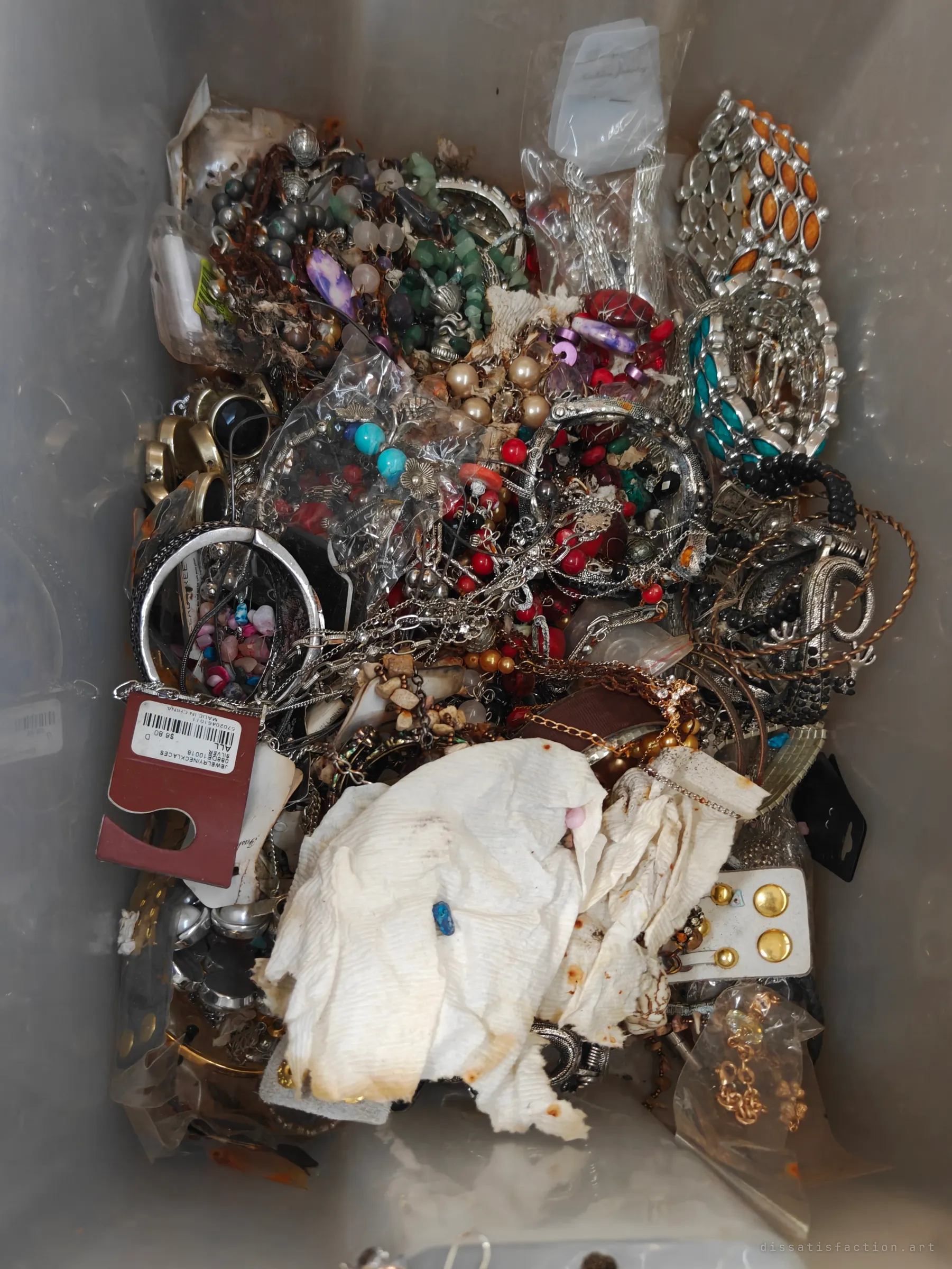 A plastic tub overflowing with necklaces, bangles and beads, some still bagged or tagged for sale, under a stained paper towel. The retail tags hint that this hoard was once merchandise.