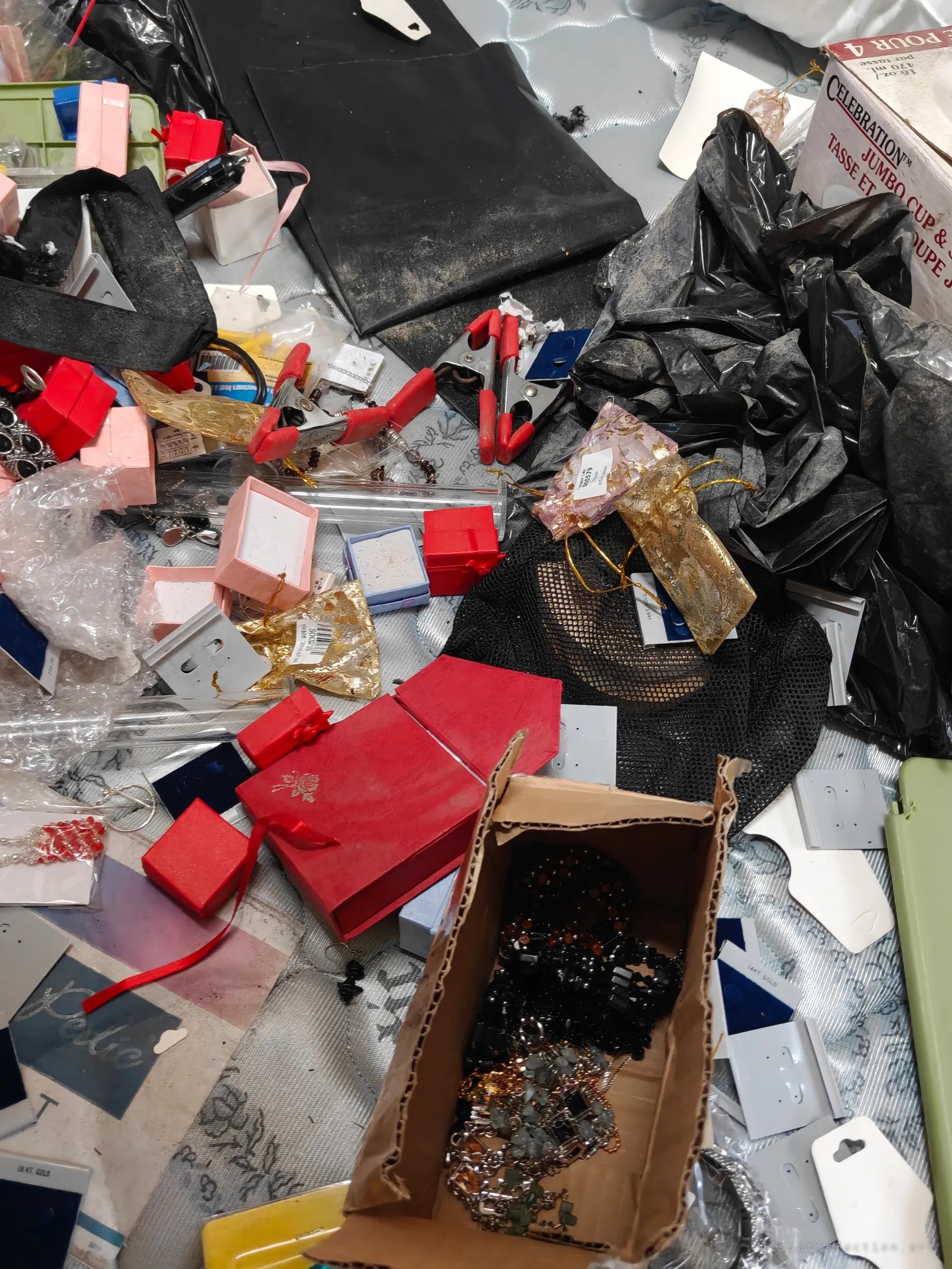 A close look at the bed's surface, strewn with red gift boxes, empty earring cards, organza pouches, clamps and a carton of beaded necklaces. The empty packaging tells the story of what was taken or sorted.