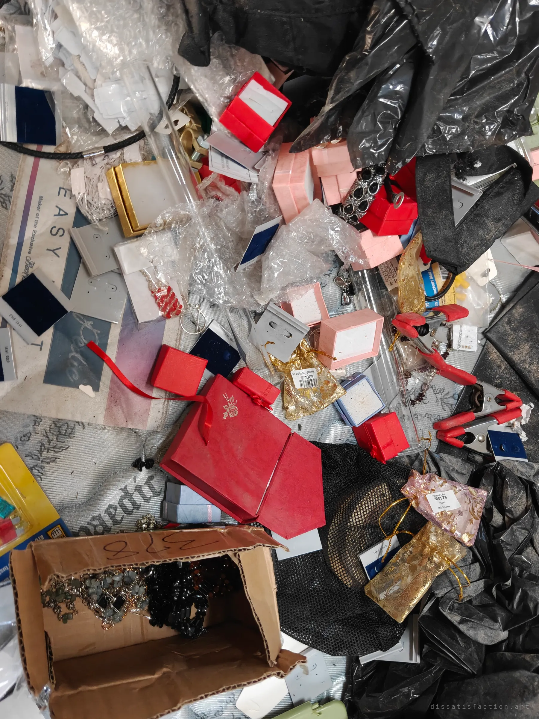 A bed buried in the stock of a jewellery seller: red and pink ring boxes, organza pouches, earring cards, bubble wrap and a cardboard box of chains. The scale of the abandoned inventory is the story of the place.