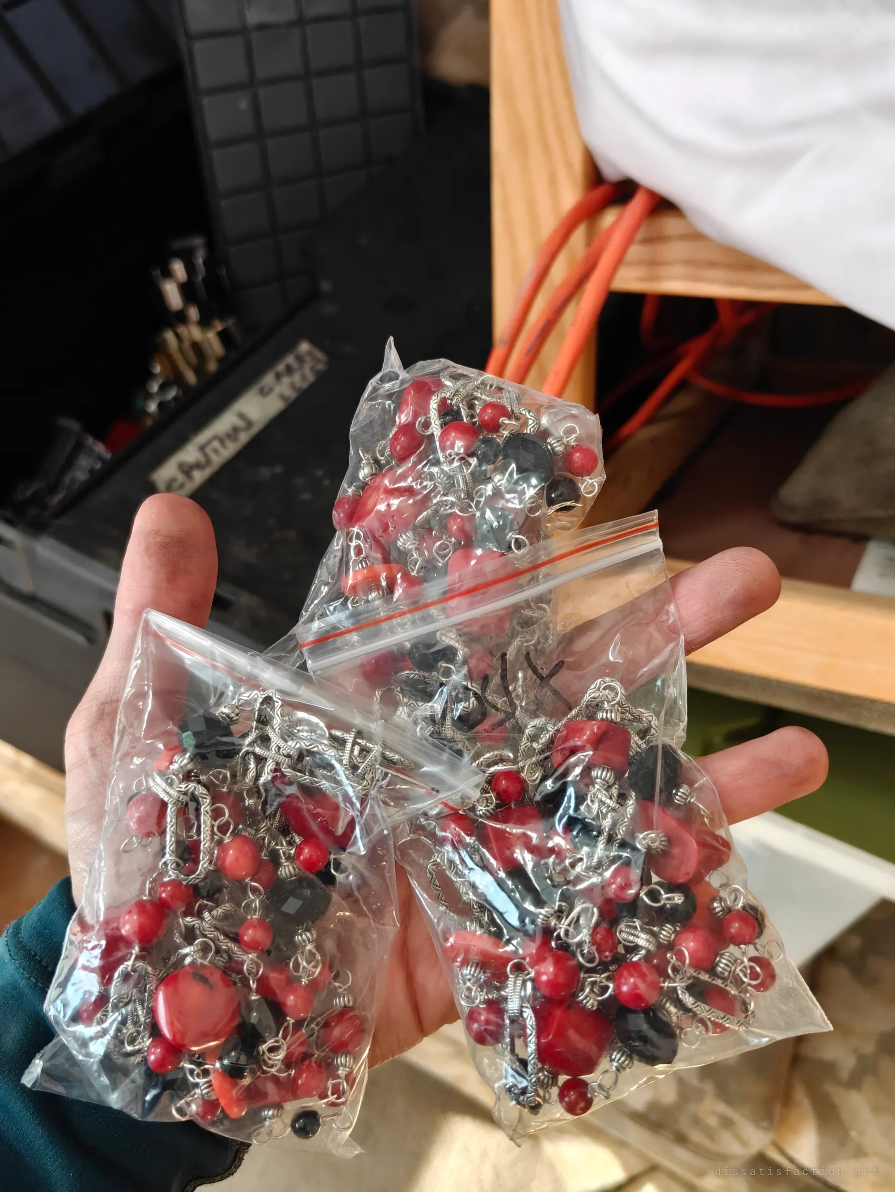 A hand holds three zip bags of red coral and black bead necklaces, pulled from storage bins beside a wooden bed frame. Sealed stock that never got sold.