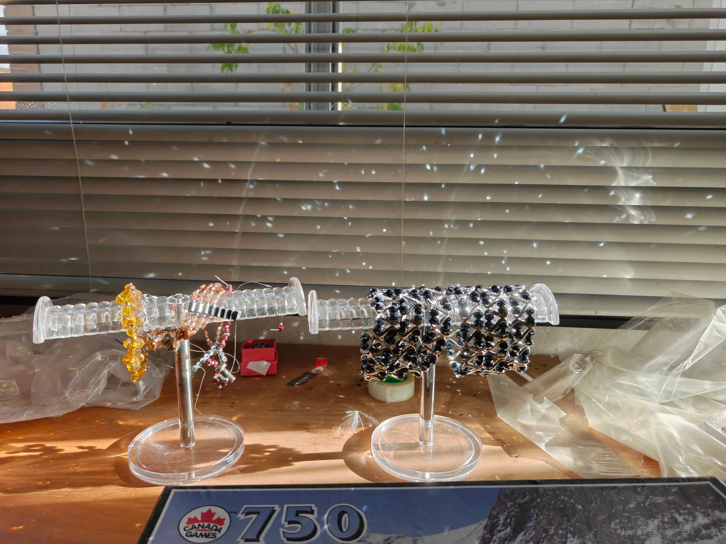 Two acrylic bracelet stands on a sunlit counter, one draped in amber and copper beads, the other in black crystal stretch bracelets, with light scattered across closed blinds. A display still set up as if waiting for buyers.