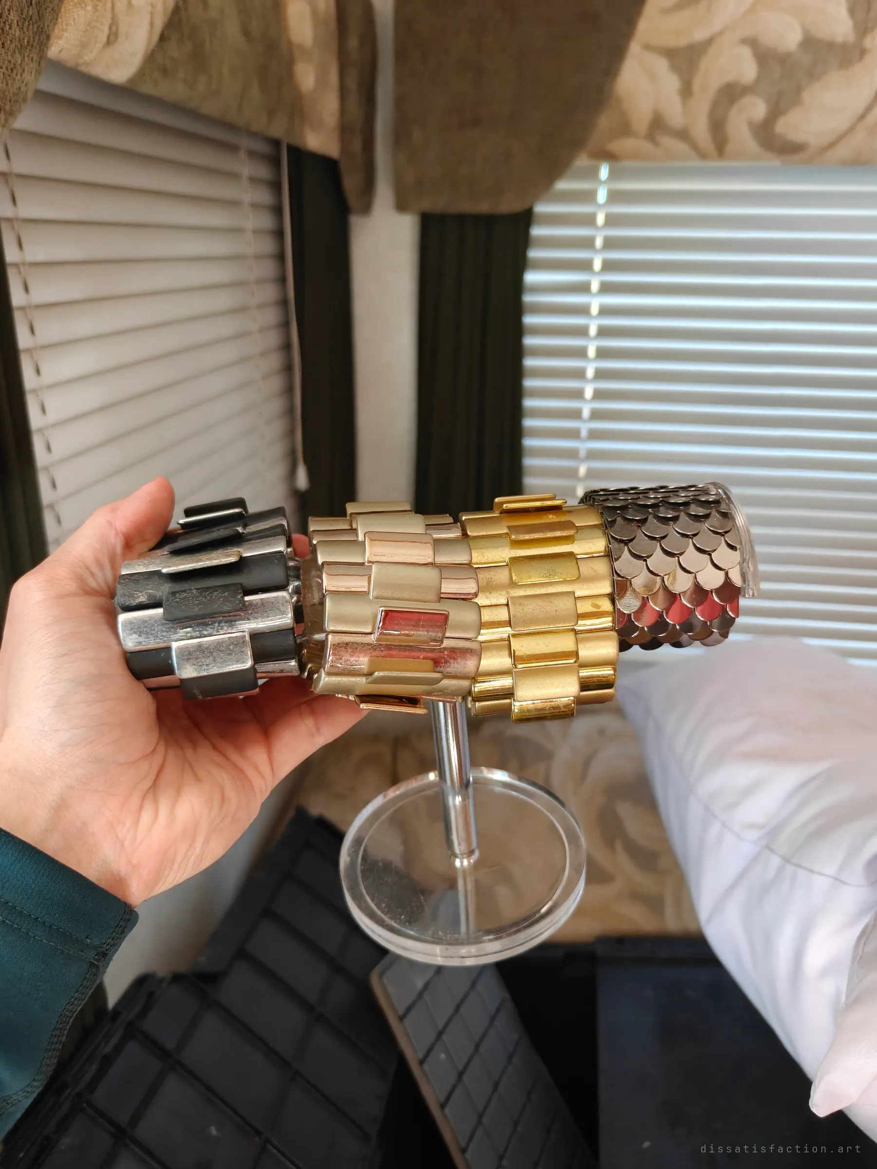 A hand tips a bracelet stand stacked with chunky link cuffs in gunmetal, champagne, gold and a scaled copper-red, in front of curtained windows. Costume pieces left behind in a cramped living space.
