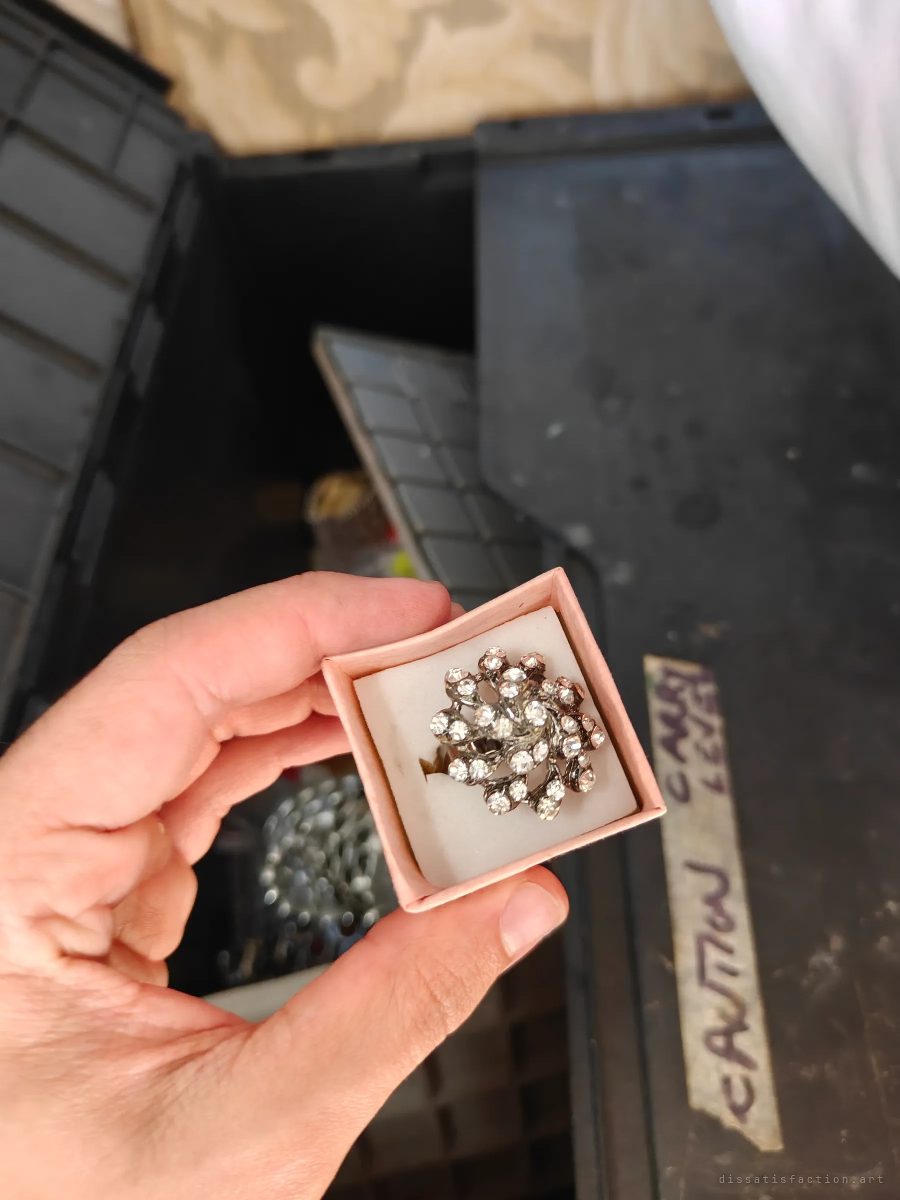 A swirl-shaped rhinestone cocktail ring in a pink box held over an open bin of more jewellery. One of many boxed pieces still waiting in the dark.