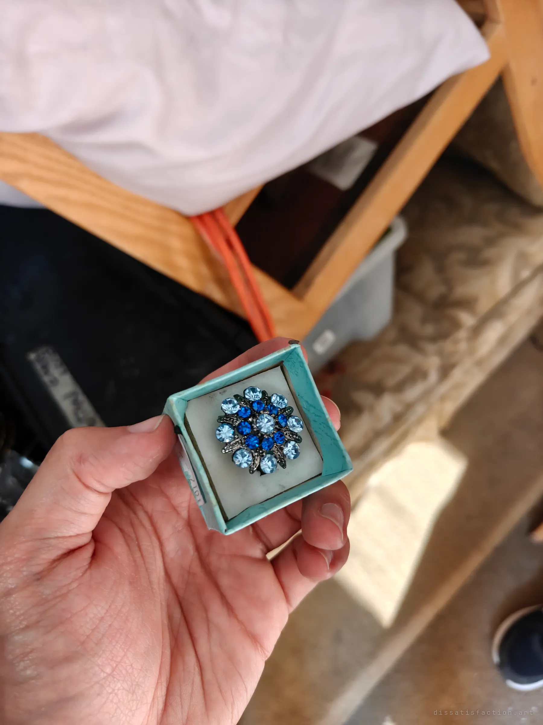 A flower-shaped ring set with light and dark blue stones in a worn turquoise box, held up over the wooden bed frame and bins. Colour among the grime.