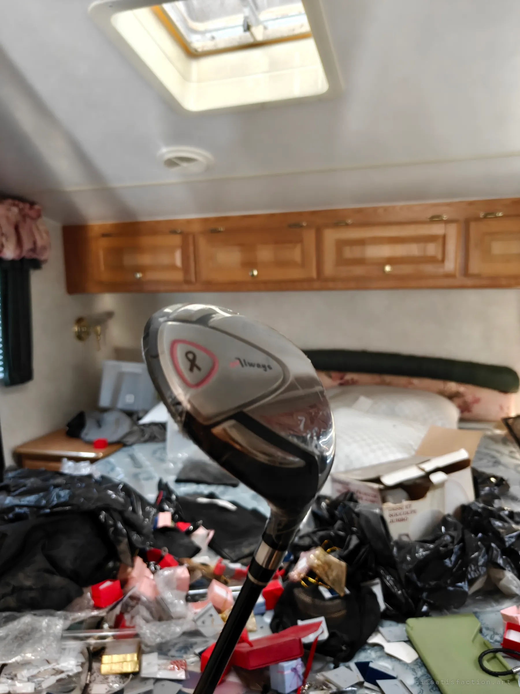 A still-wrapped golf club held up in the bedroom of a camper, the bed behind it drowning in jewellery boxes and black bags under oak cabinets and a roof vent. It shows how tight and overloaded the space is.