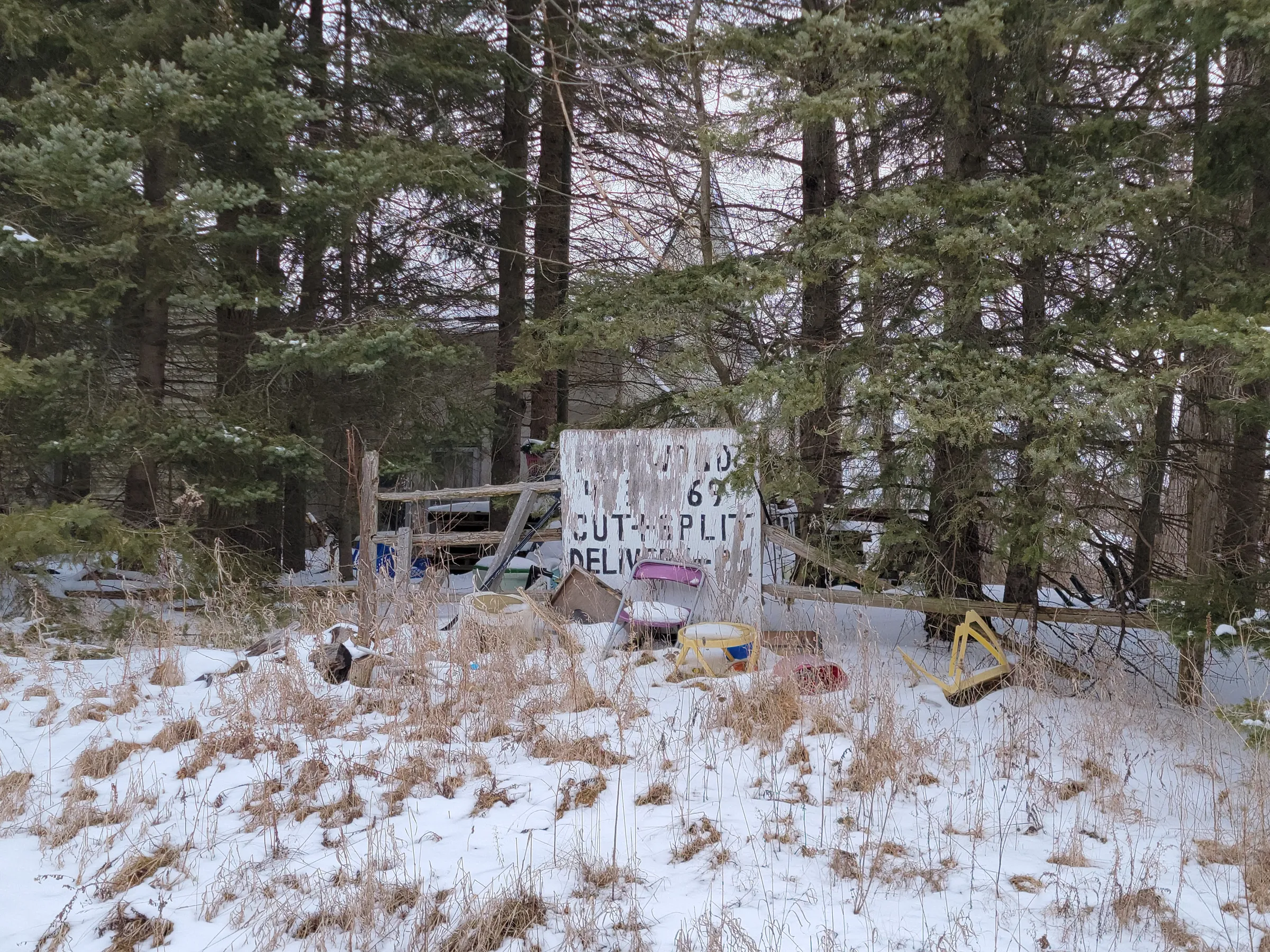 A peeling hand-painted firewood sign propped among snow, dead grass and junk: a folding chair, a toy walker, buckets. The yard is already filling with the household's overflow.