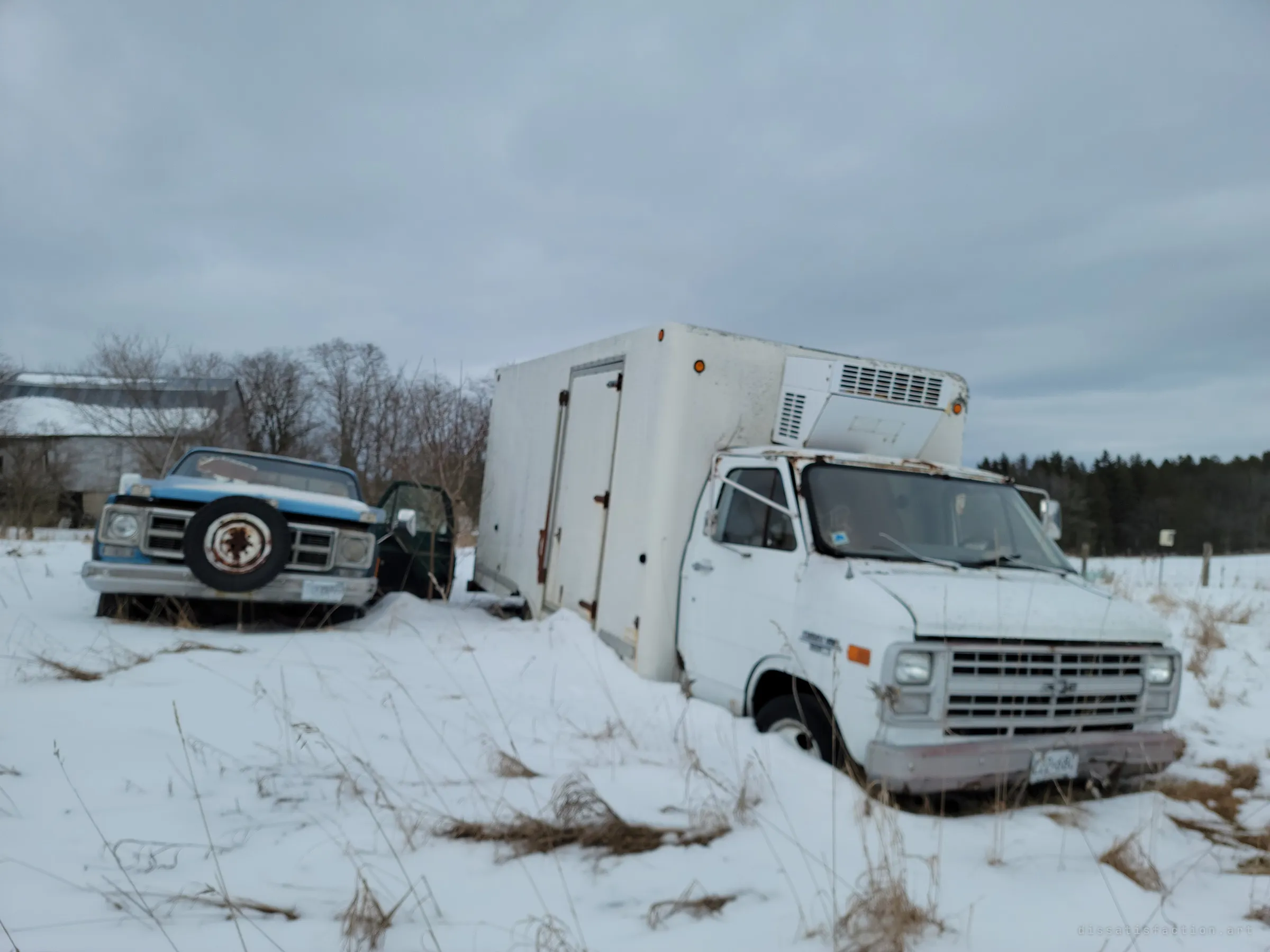 An old blue pickup and a white refrigerated box truck sinking into a snowy field, a barn roof behind them. The vehicles were left where they last stopped.