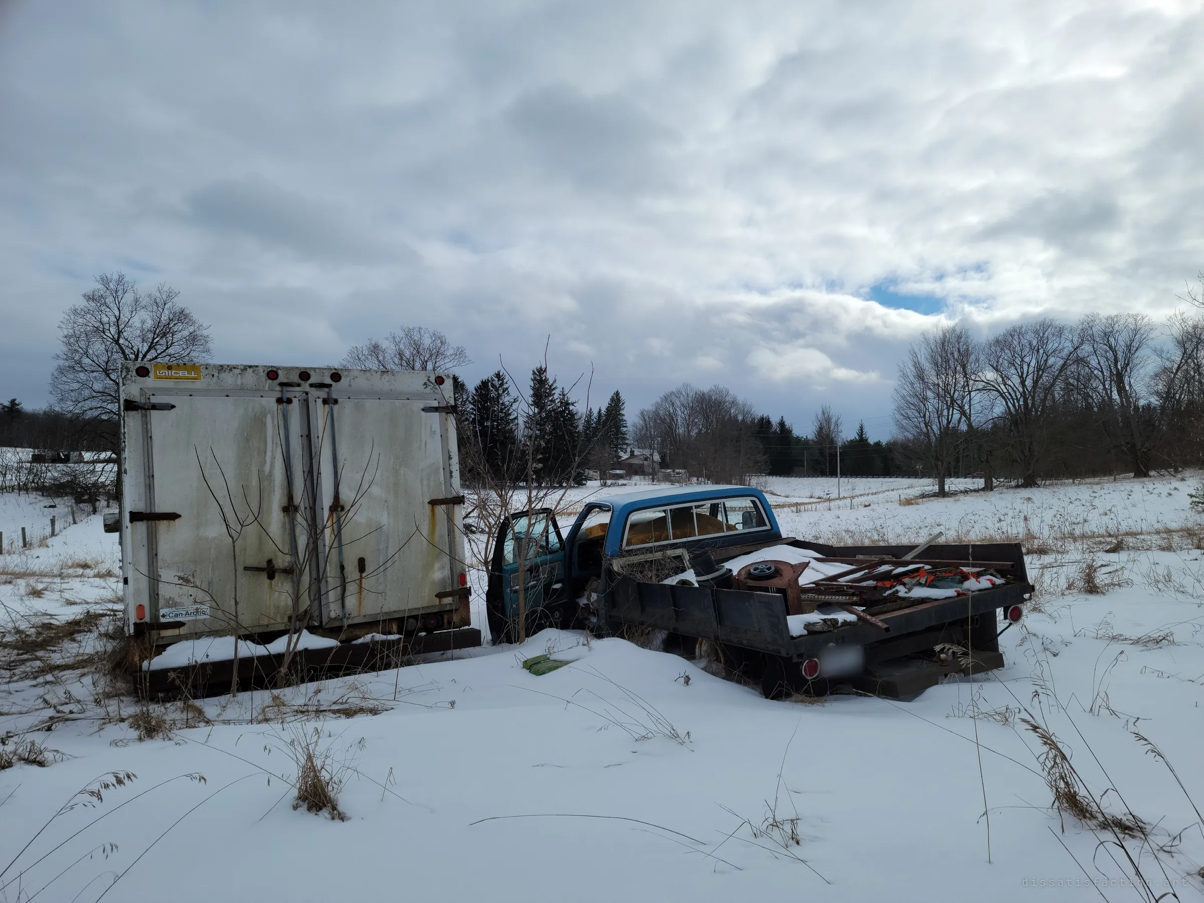 The back of the white box truck beside a blue flatbed pickup heaped with scrap metal, both sinking into a snowy field under a broken sky. The truck's rear doors still open onto whatever was stored inside.