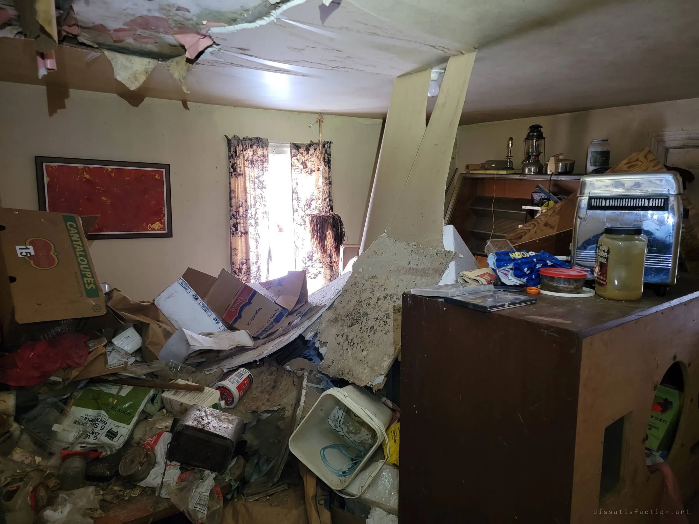 Inside the house the ceiling has collapsed onto waist-high piles of boxes, food containers and rubbish, light coming through a curtained window past a red painting and an old toaster.