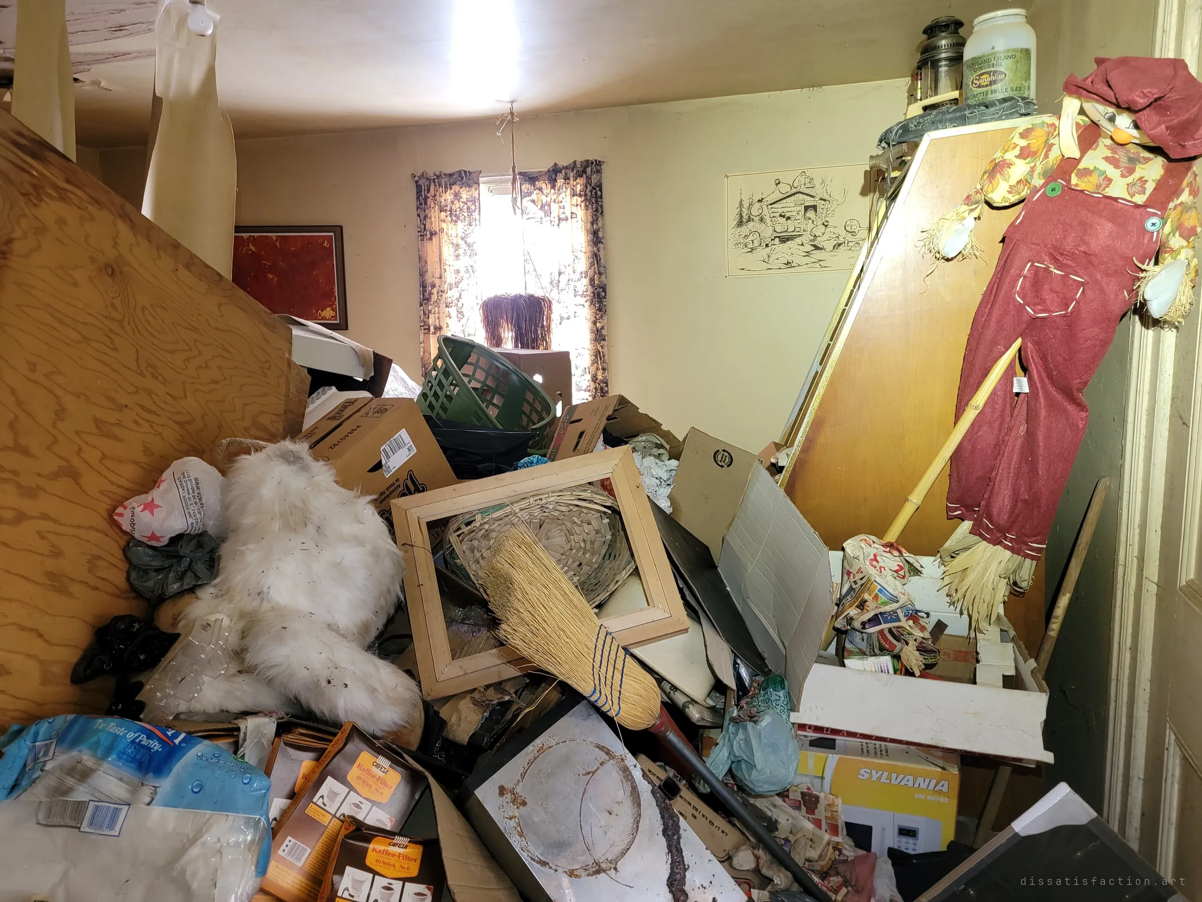 The same room from closer in: a scarecrow doll, a white teddy bear, a broom and picture frames heaped against a wall with a cartoon print of a log cabin. Decades of belongings piled to the ceiling.