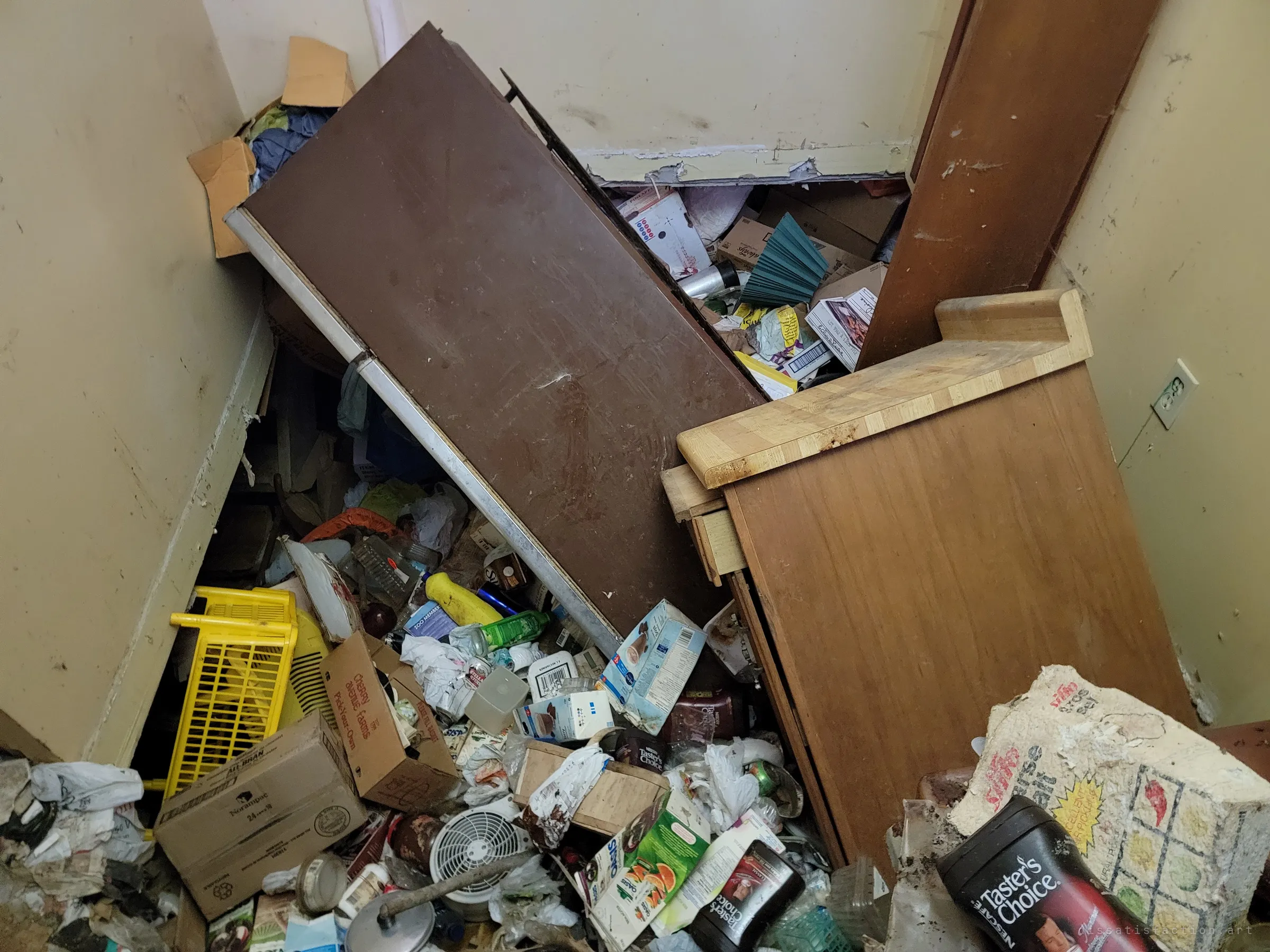 Looking down into a narrow corner where a fallen cabinet and a wooden counter lie across a drift of boxes, bottles and coffee jars. Nothing here has been cleared in years.