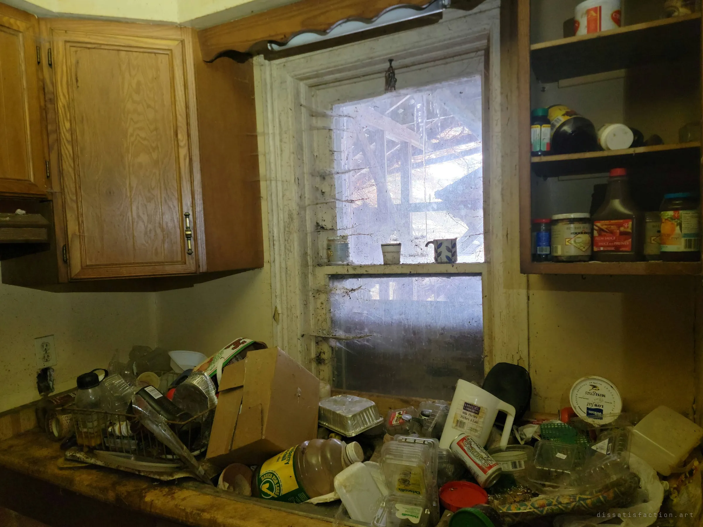 A kitchen counter buried under empty containers and packaging beneath a cobweb-draped window. Food is still in the open cupboard, as if the owner simply stopped.