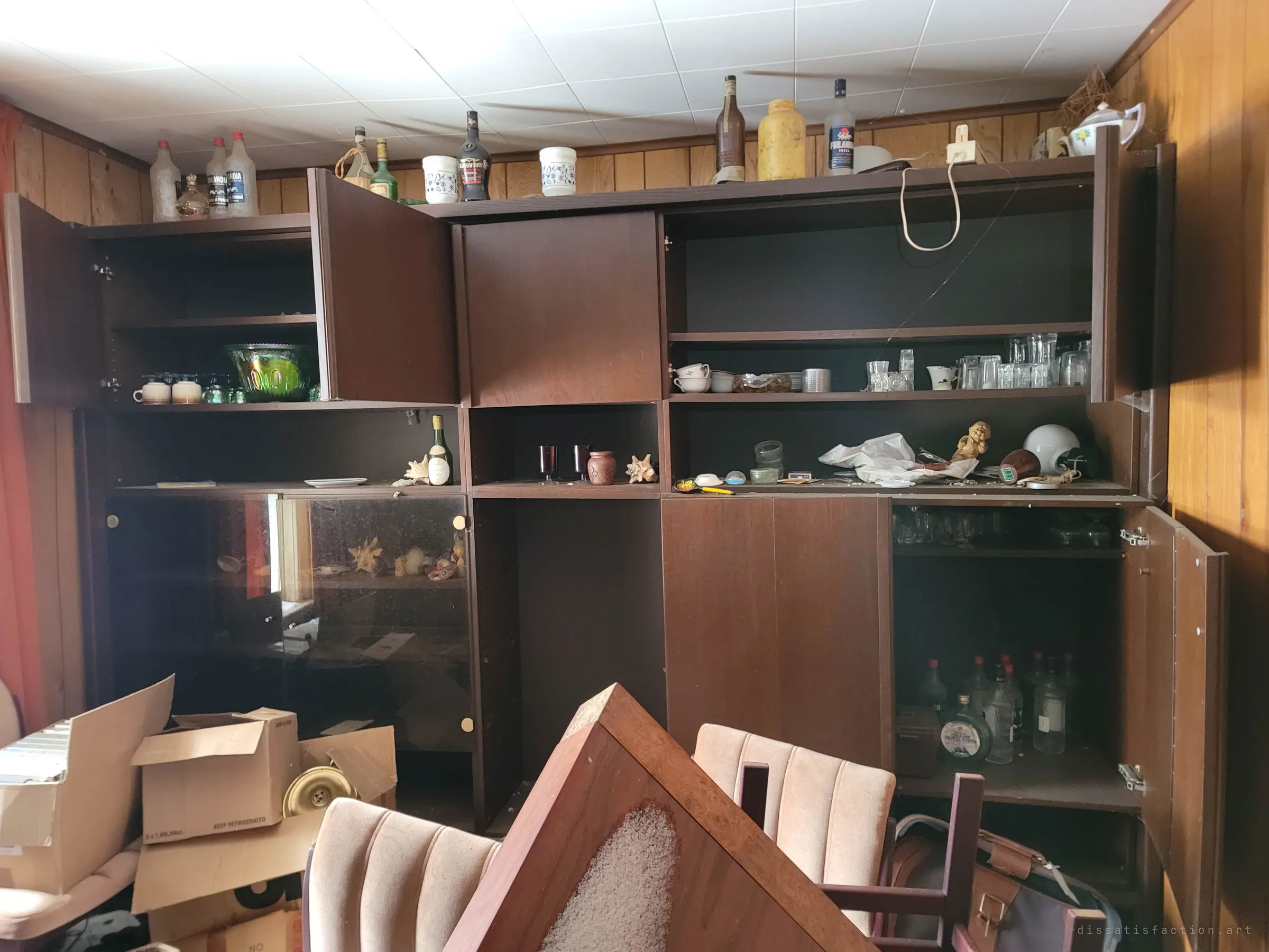 A wood-panelled dining room wall unit with open cabinets of glassware and liquor bottles, an overturned table and chairs piled in front. The room has been half dismantled but never emptied.