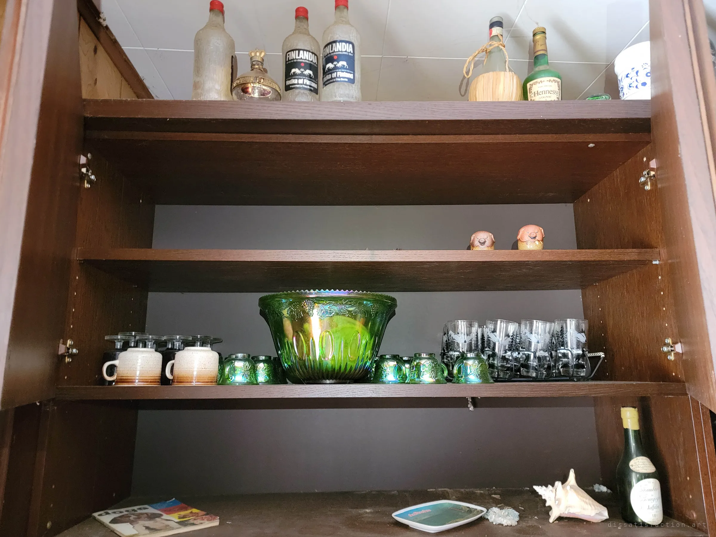Close-up of an open cabinet holding an iridescent green punch bowl set, mugs and old vodka bottles. Carefully kept tableware left behind in the dust.