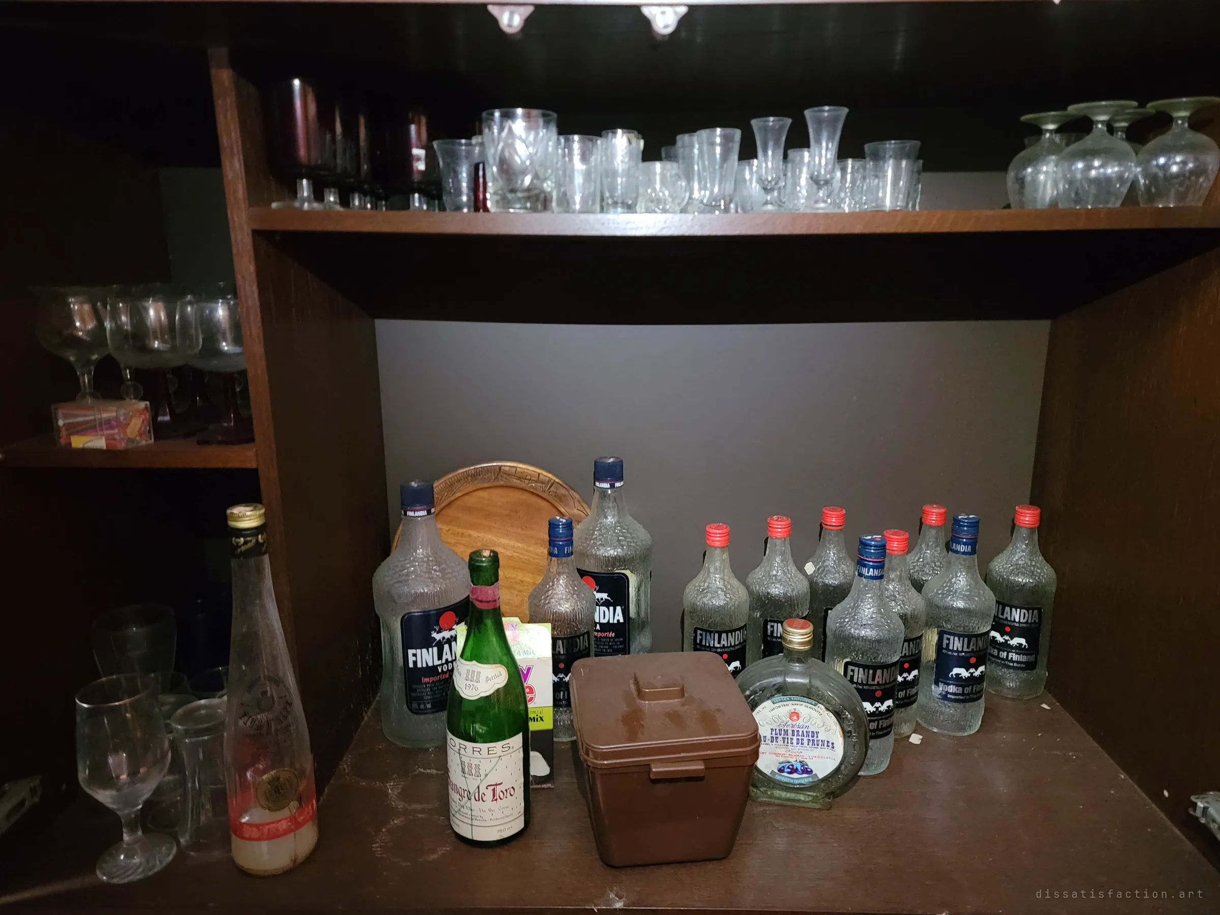 A dark liquor cabinet crowded with rows of empty vodka bottles, a vintage wine and an ice bucket under shelves of glasses. Decades of collected bottles never thrown away.