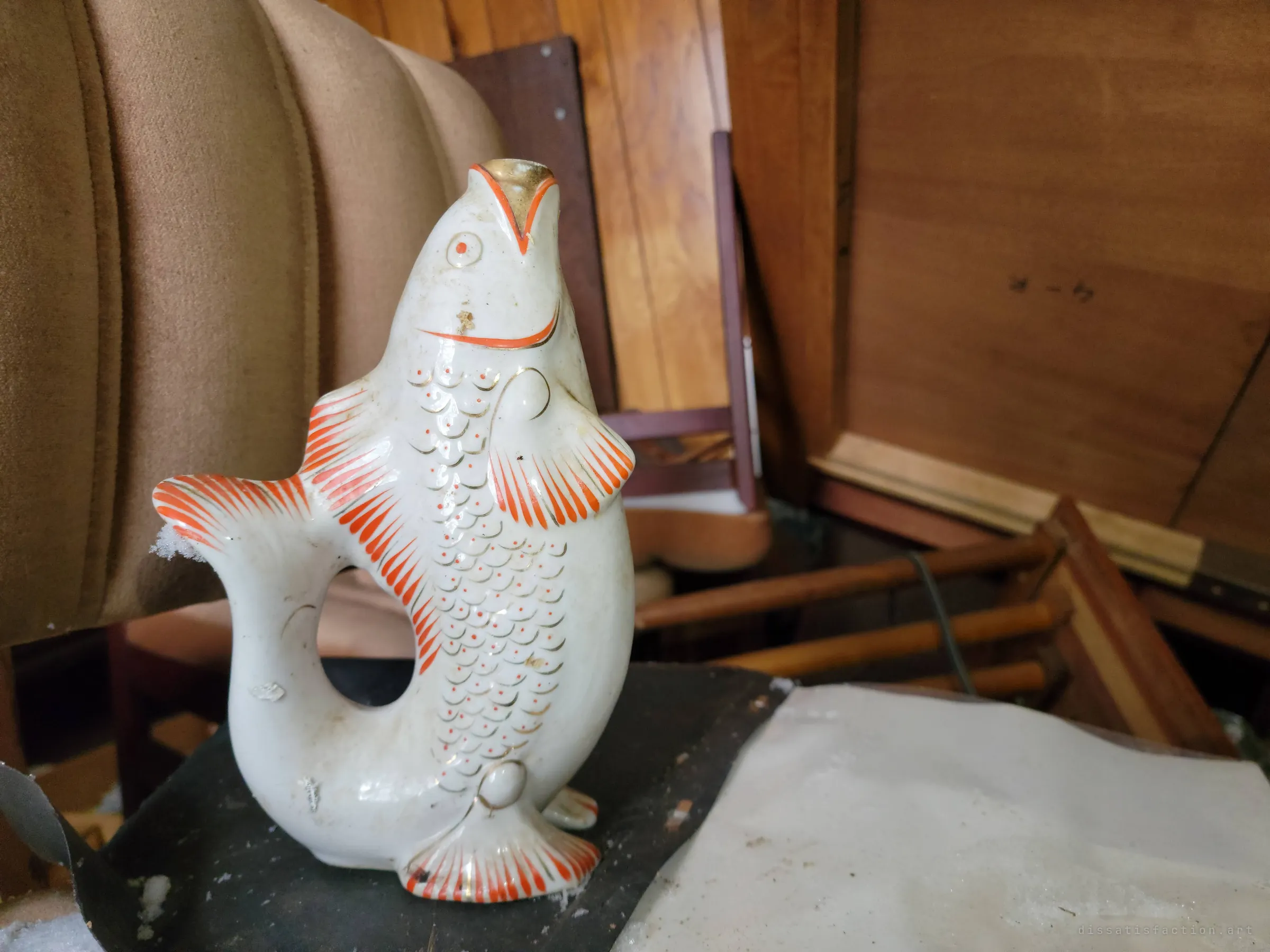 A hand-painted porcelain fish decanter standing on a chair among stacked furniture. A small ornament that survived the chaos intact.