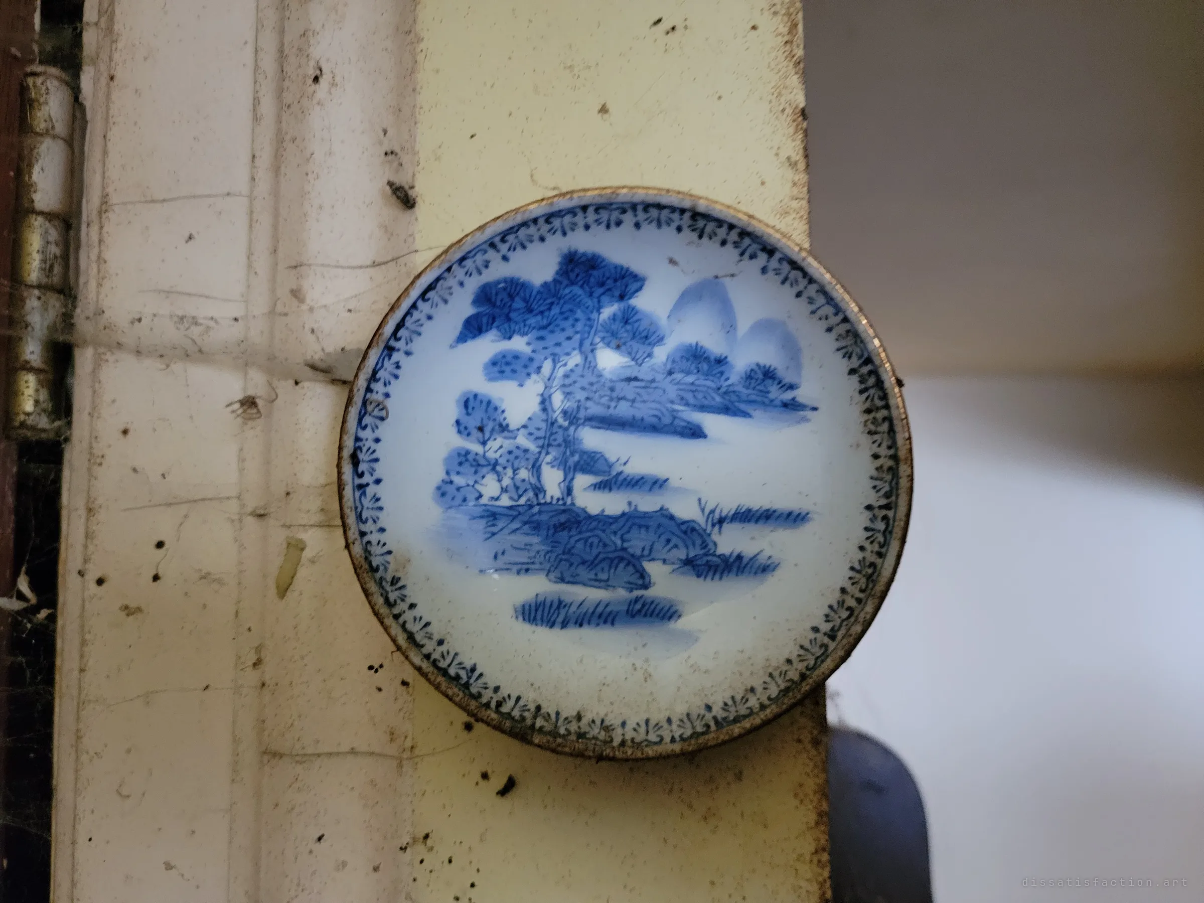 A small blue-and-white painted dish of a tree-and-mountain landscape set into a dirty door frame. A delicate detail amid grime and cobwebs.