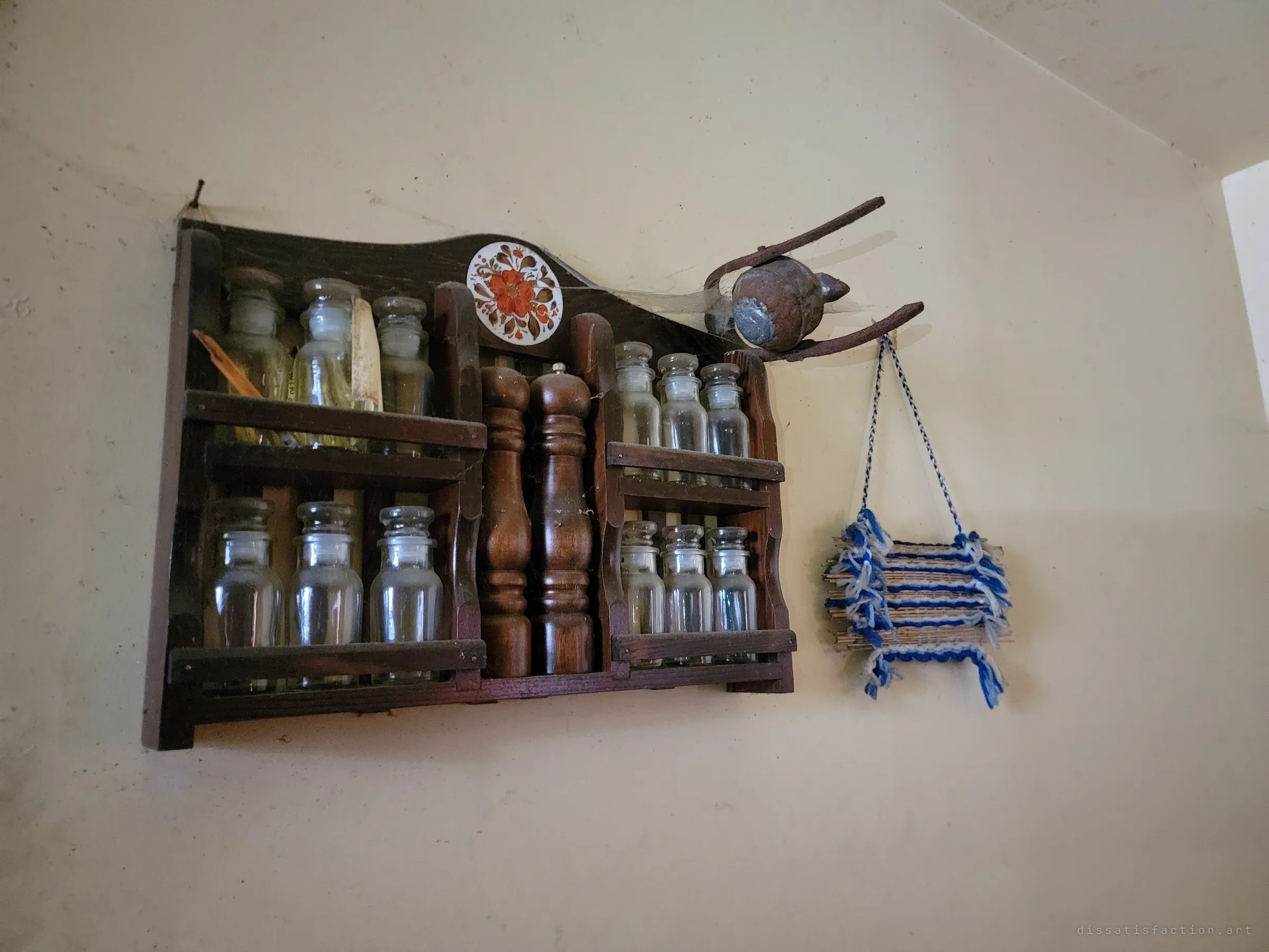 A wooden spice rack of empty glass jars and pepper mills hanging on a bare kitchen wall, draped in cobwebs, beside a woven hanging. Domestic order left to gather dust.