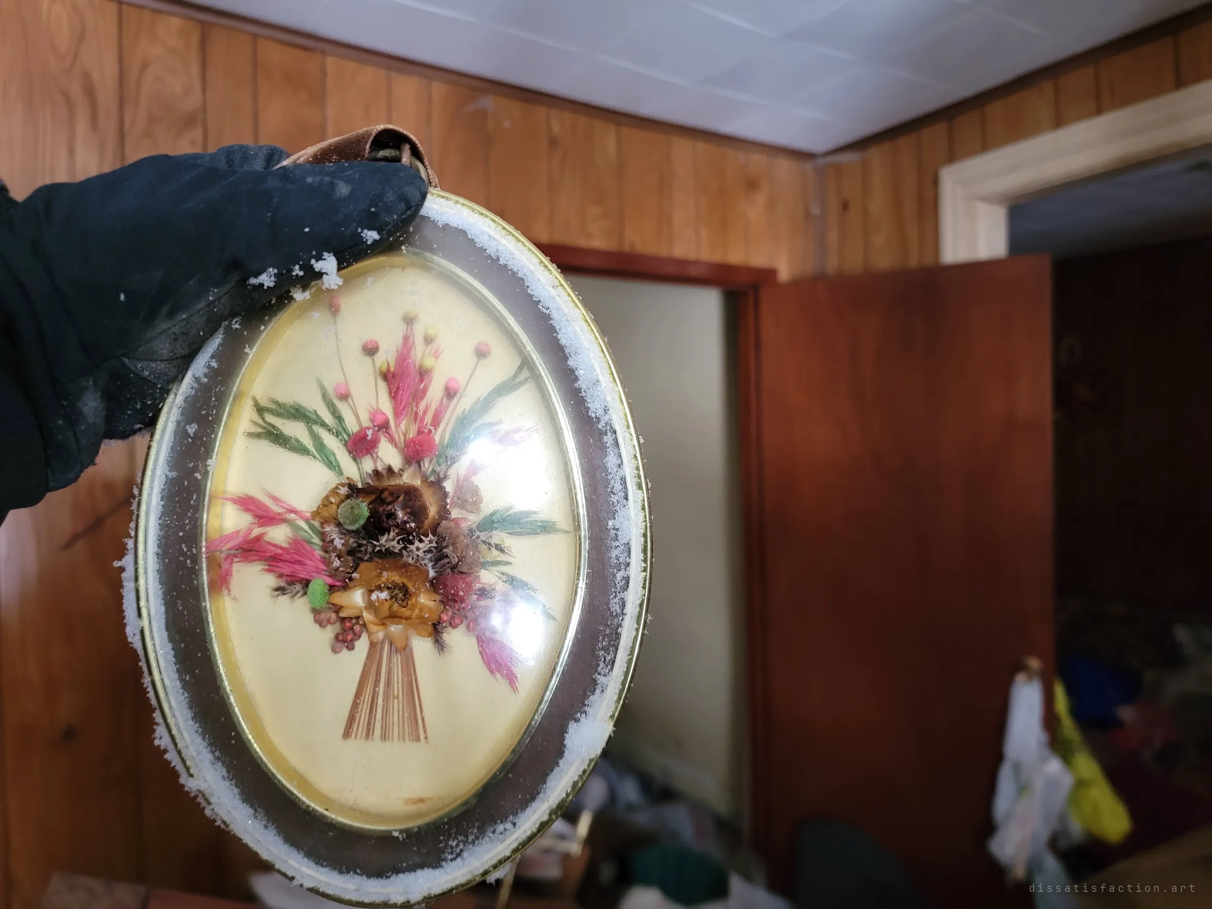 A gloved hand holds up a domed frame of dried flowers dusted with snow, panelled rooms blurred behind. Snow indoors shows how open to the weather the house has become.