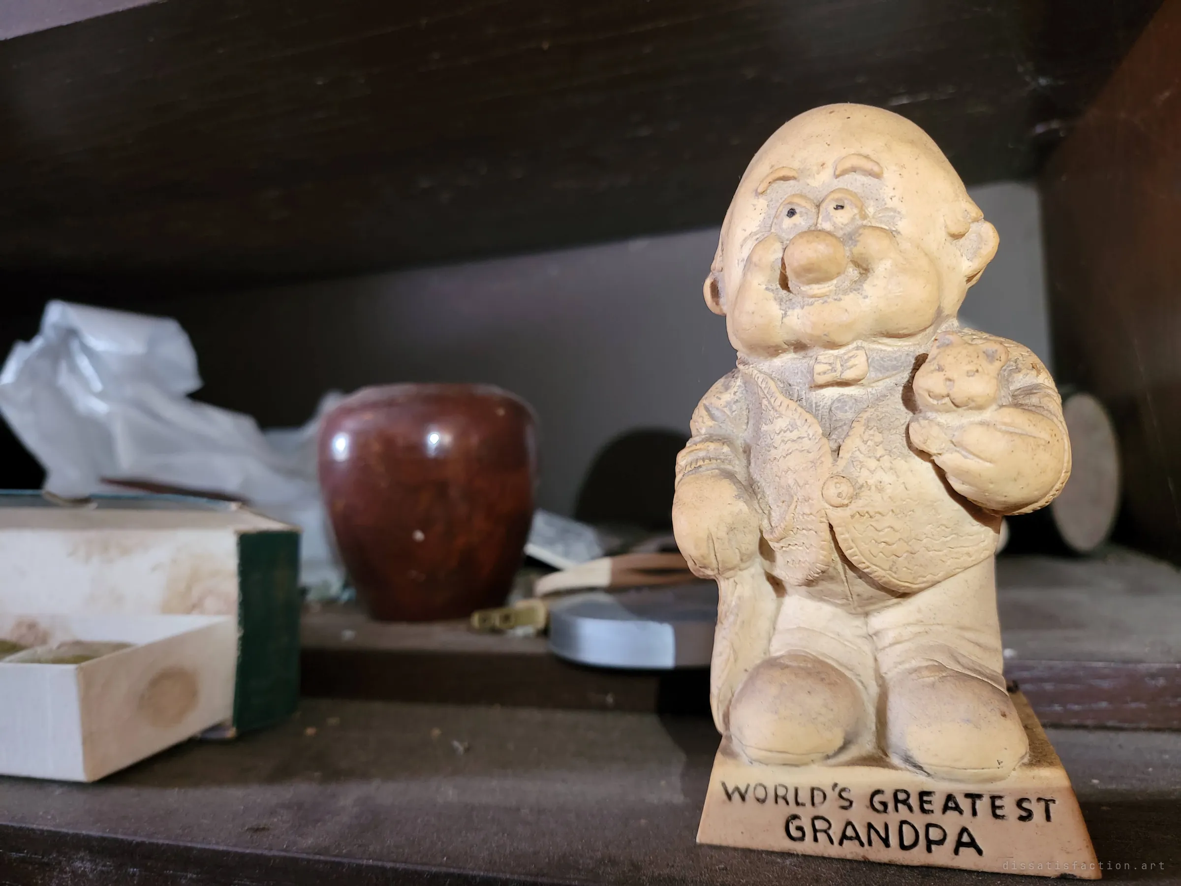 A dusty novelty figurine of a grandfather holding a teddy bear on a cabinet shelf. A family keepsake left behind with everything else.