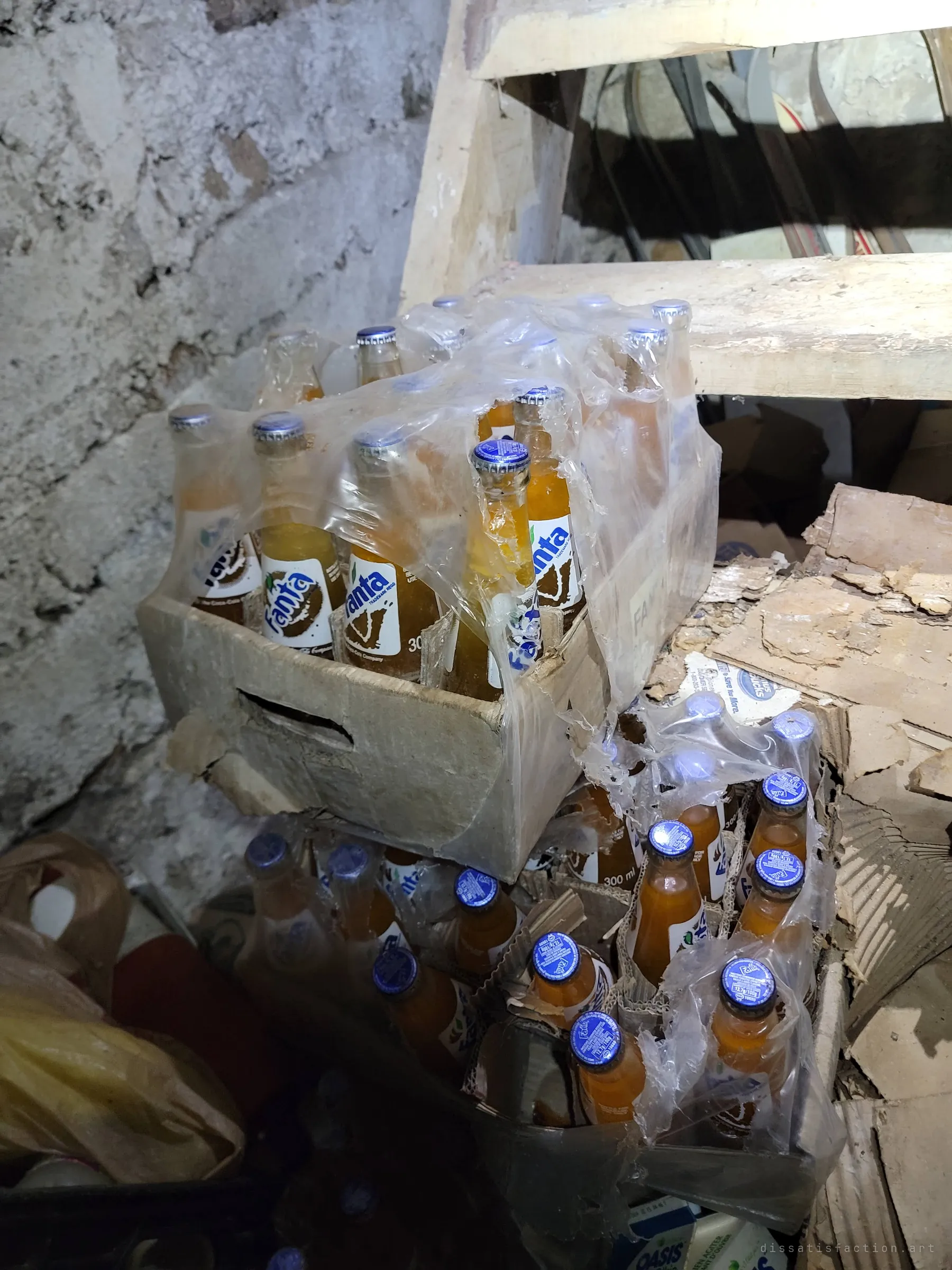 Shrink-wrapped cases of old glass Fanta bottles sit unopened beneath a rough wooden basement staircase against a whitewashed stone wall.