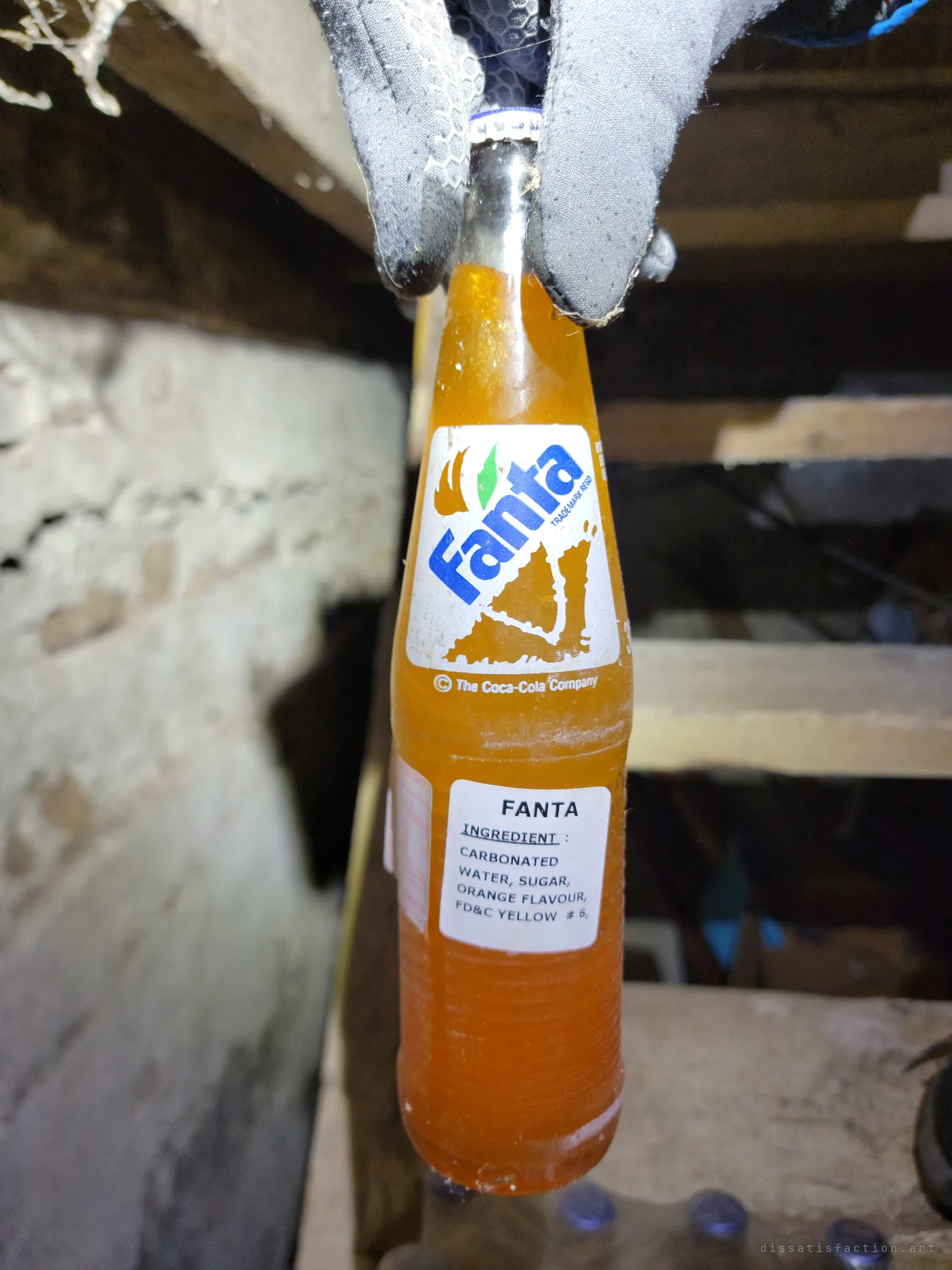 A gloved hand dangles one of the vintage Fanta bottles, its orange soda still sealed, in front of the basement stairs and stone foundation.