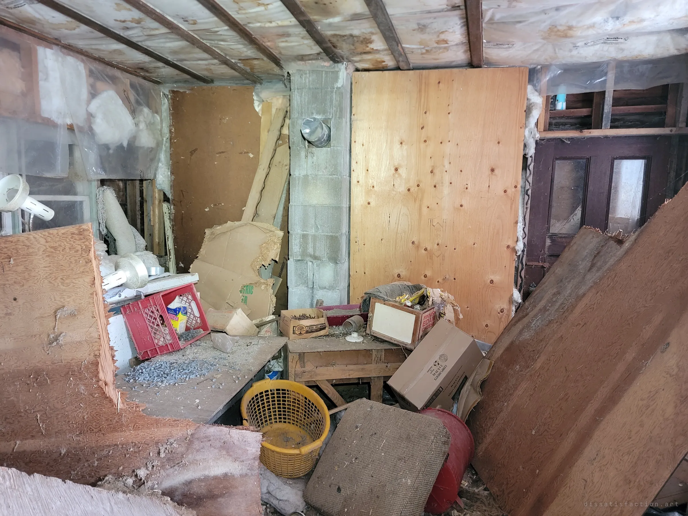 A half-gutted back room with exposed joists and insulation, a cinder-block chimney, plywood sheets and a workbench scattered with nails and crates.