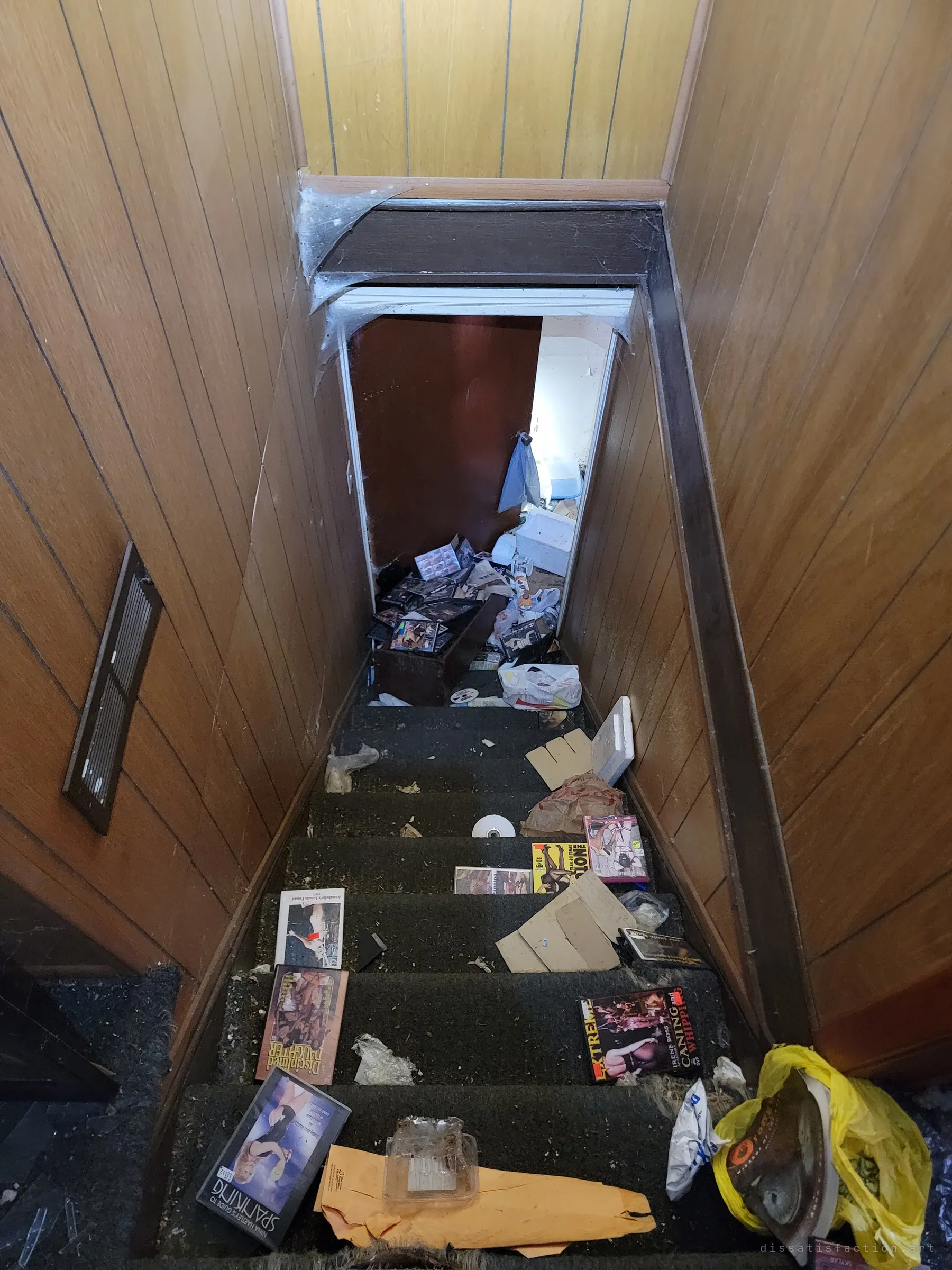 Looking down a narrow wood-panelled staircase strewn with adult DVDs and trash toward a cluttered landing below.
