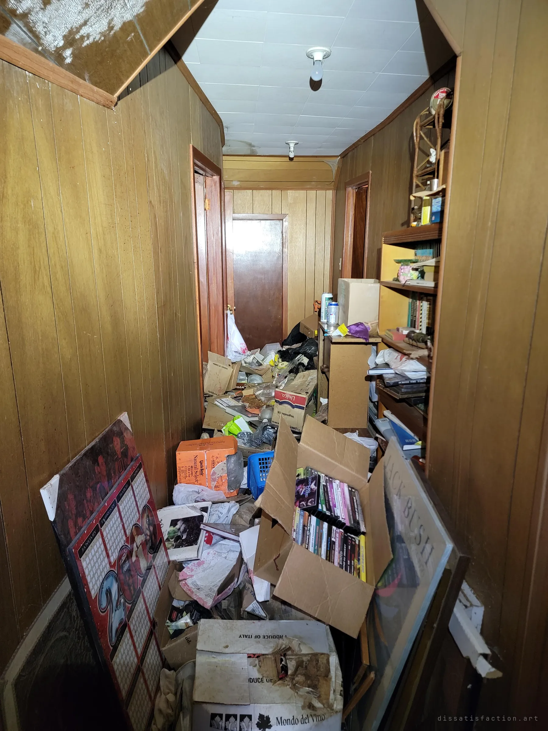 An upstairs hallway in wood panelling buried knee-deep in boxes, DVDs, an old wall calendar and bags, with closed doors waiting at the end.