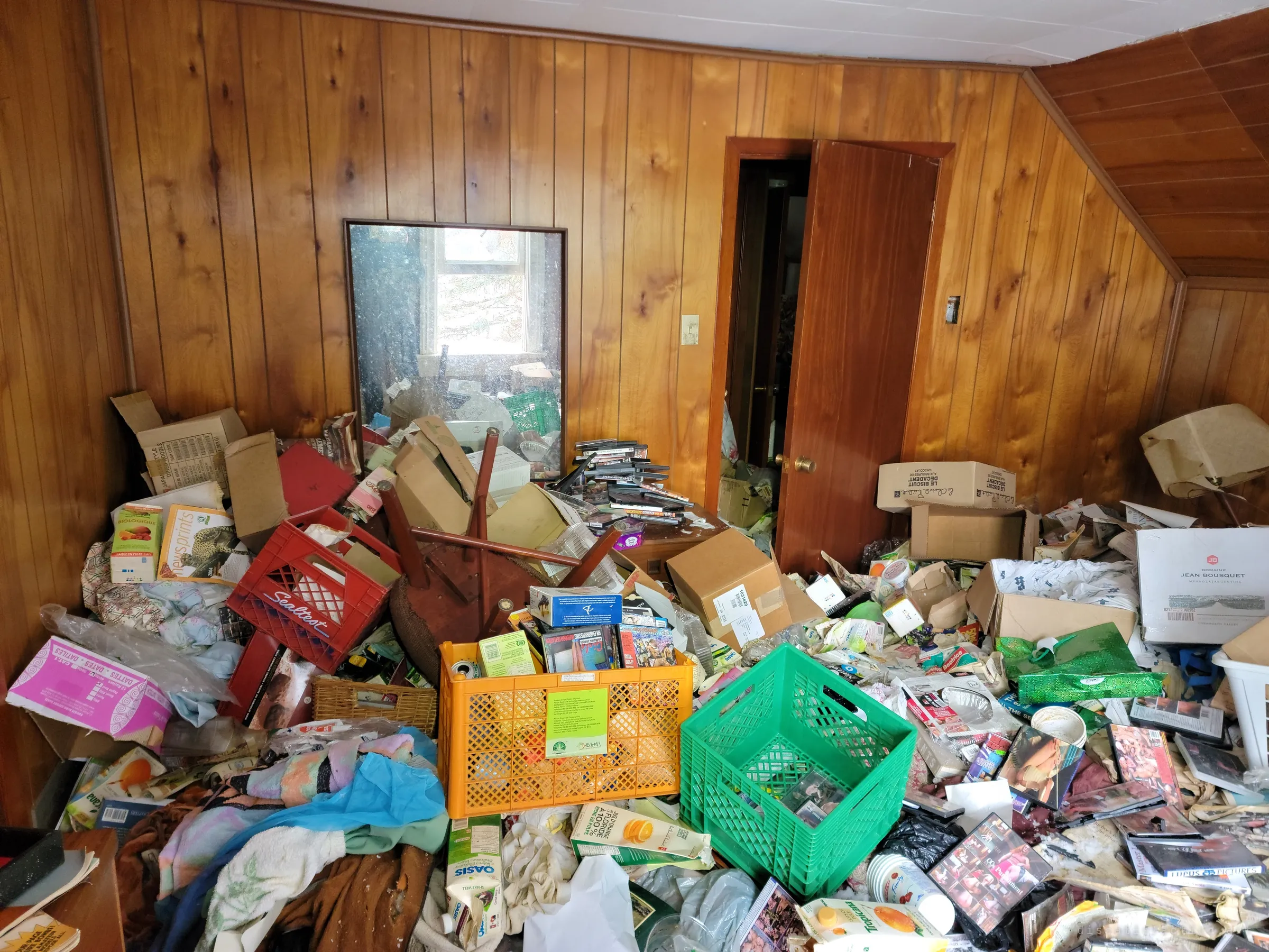 An attic bedroom in knotty pine panelling submerged under crates, boxes, clothes and scattered DVDs, a mirror on the far wall catching the window light.