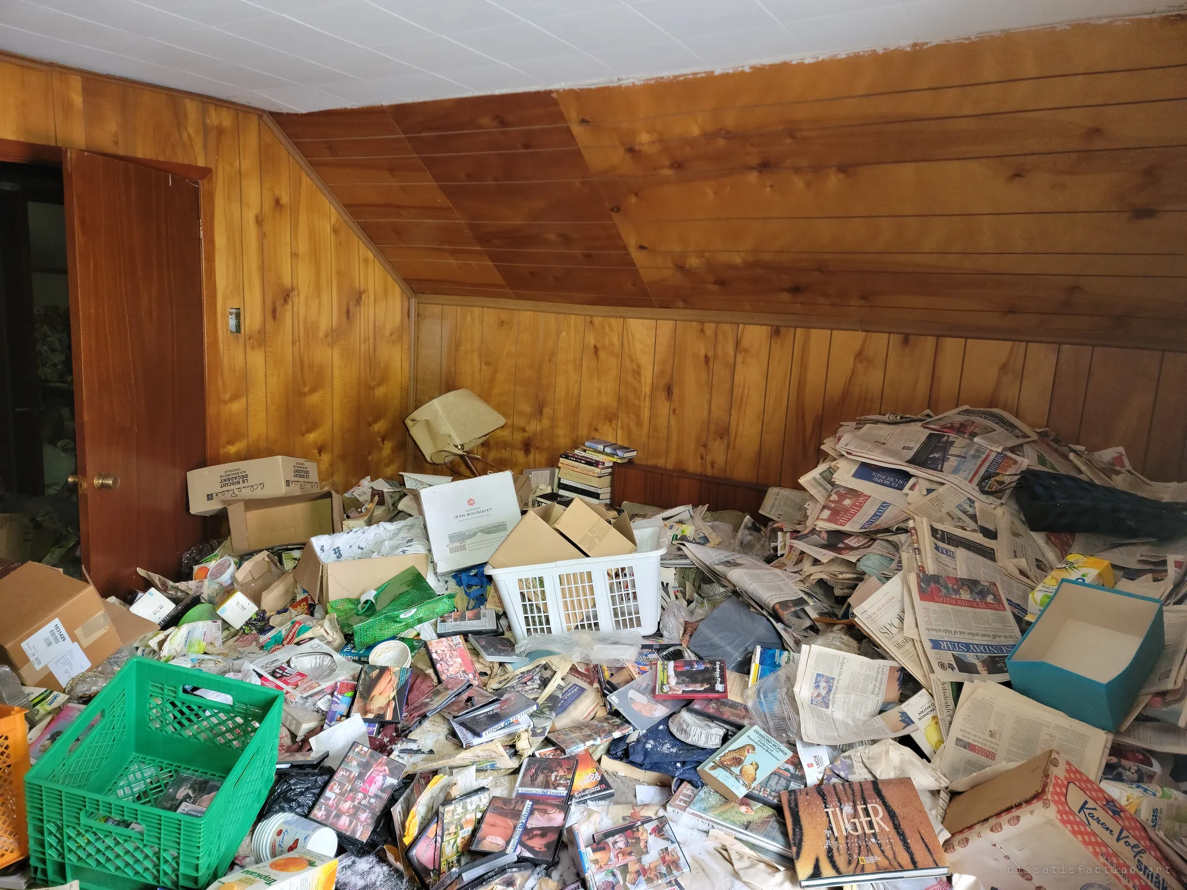 An attic bedroom under sloped pine panelling, its floor buried knee-deep in newspapers, DVDs, crates and boxes. The sheer volume of hoarded paper sets the scale of the house's collapse.