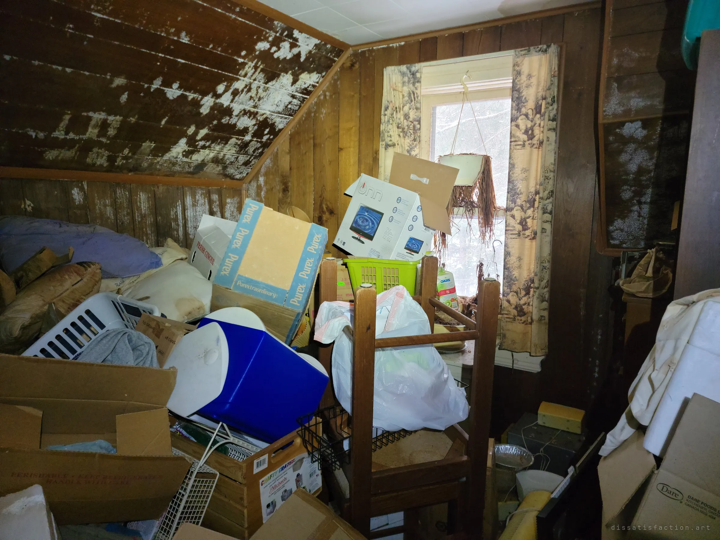 A small attic room with mould-streaked panelling, packed with coolers, boxed electronics, laundry baskets and an upturned chair beneath a curtained window. Light from the single window shows how far the clutter reaches.