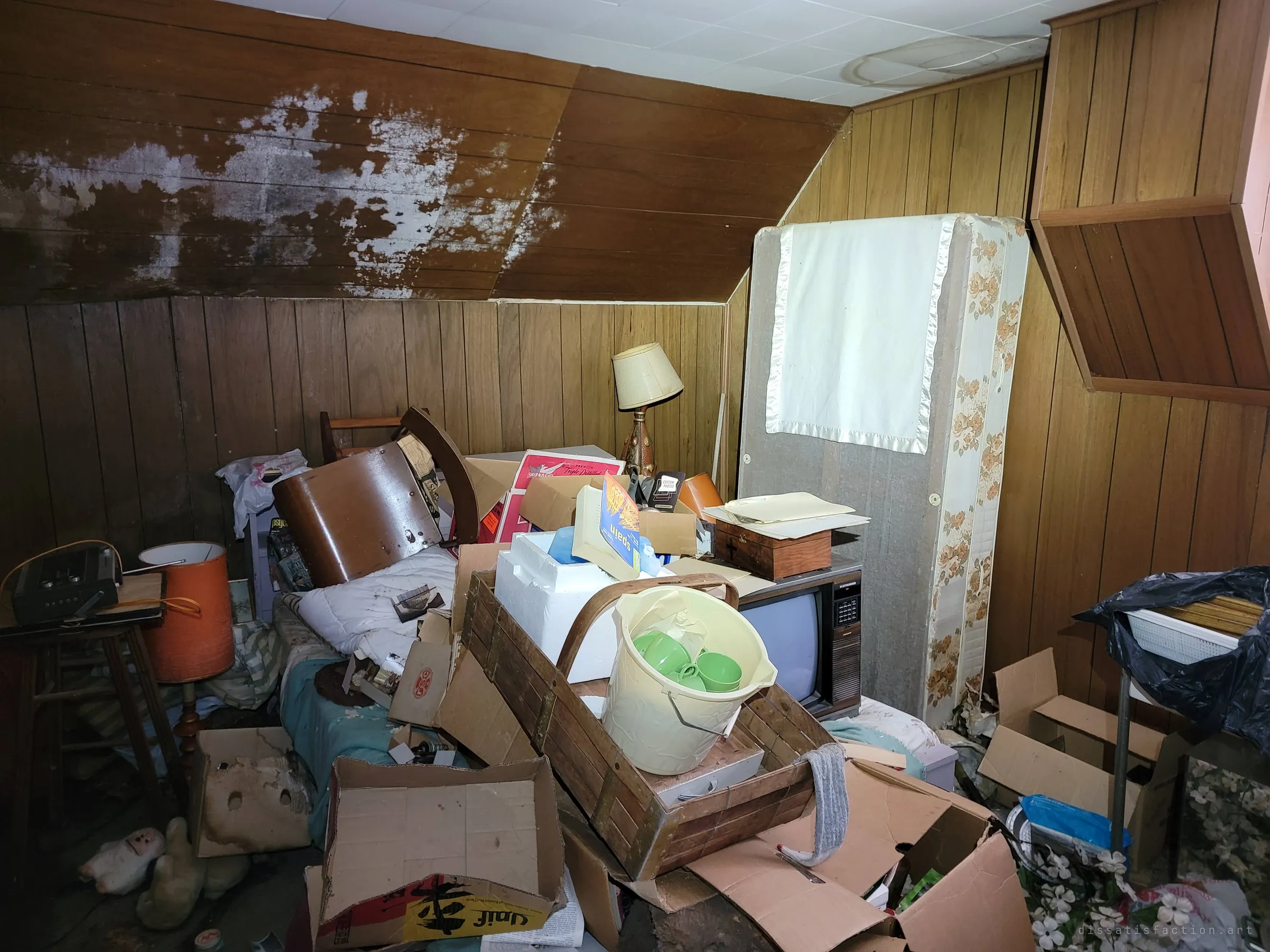 A mould-bleached sloping ceiling over a bedroom crowded with a bed, an old tube TV, lamps and a mattress stood on end. The bed is still made underneath the piles.