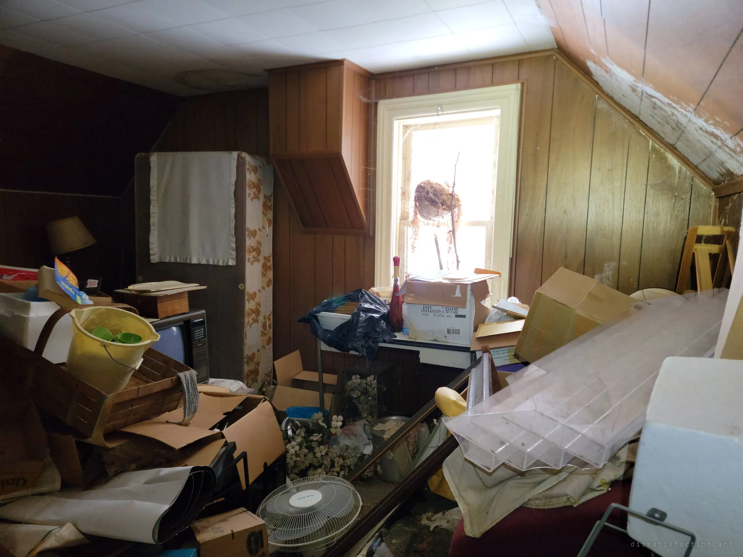 The same attic bedroom seen toward its bright window, where a dead hanging plant still hangs over boxes, a fan and sheets of clear plastic. The window is the only open space left in the room.