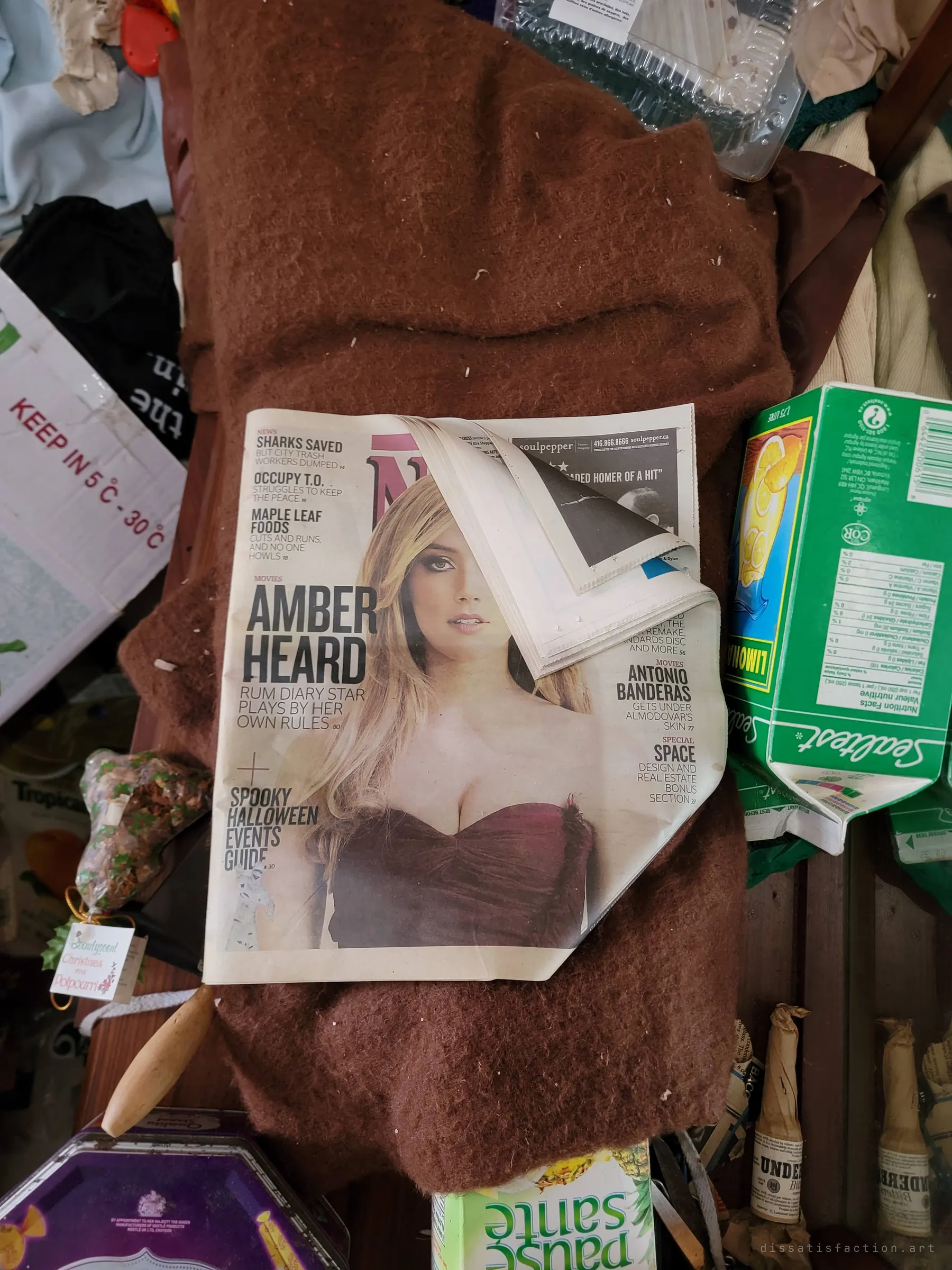 A free city weekly with a film star on the cover, lying on a brown blanket among cartons and tins. Old magazines like this help date when life in the house stopped.