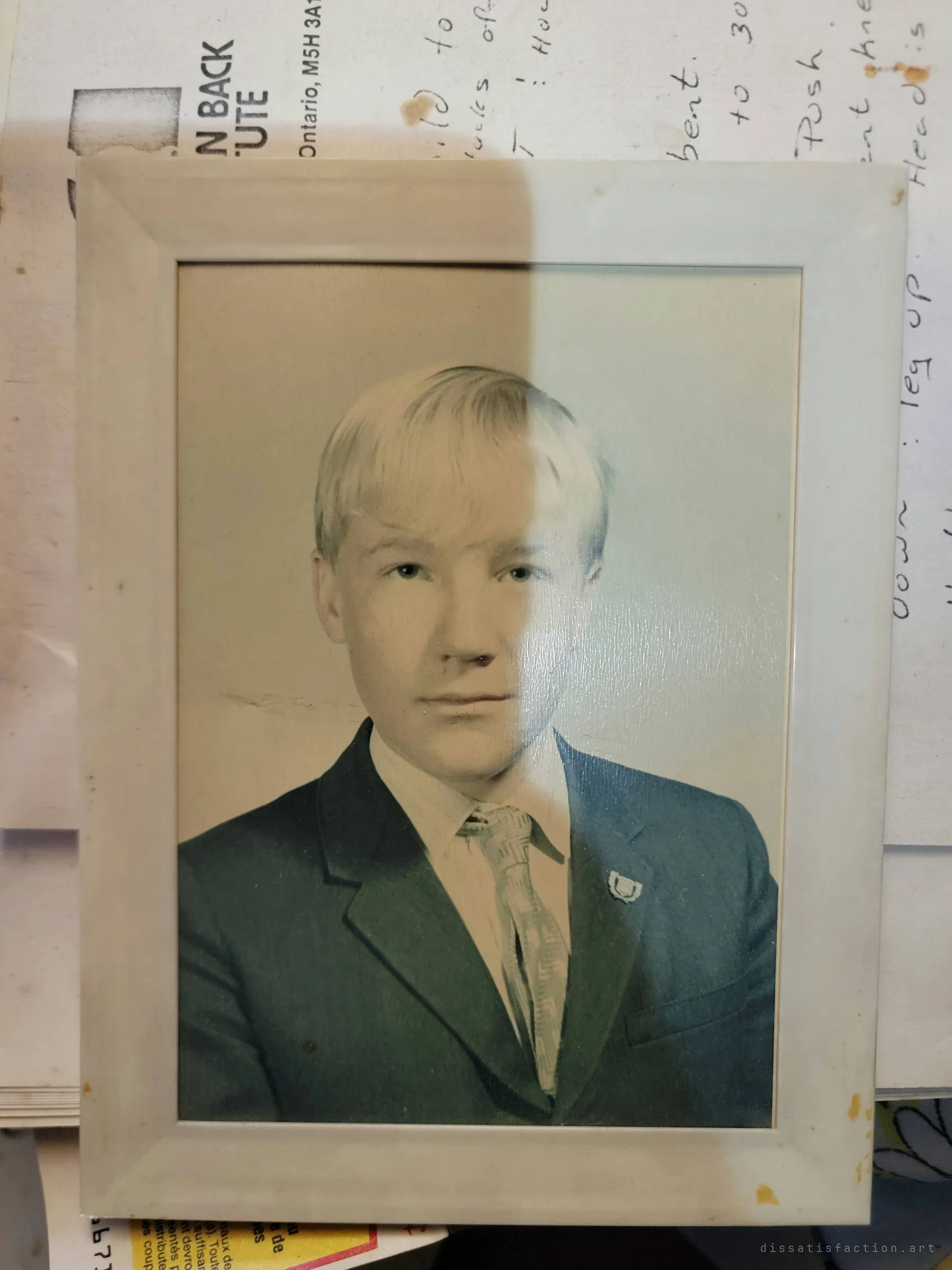 A framed, hand-tinted school portrait of a blond teenager in a suit, resting on handwritten exercise notes. One of many personal keepsakes abandoned with the house.