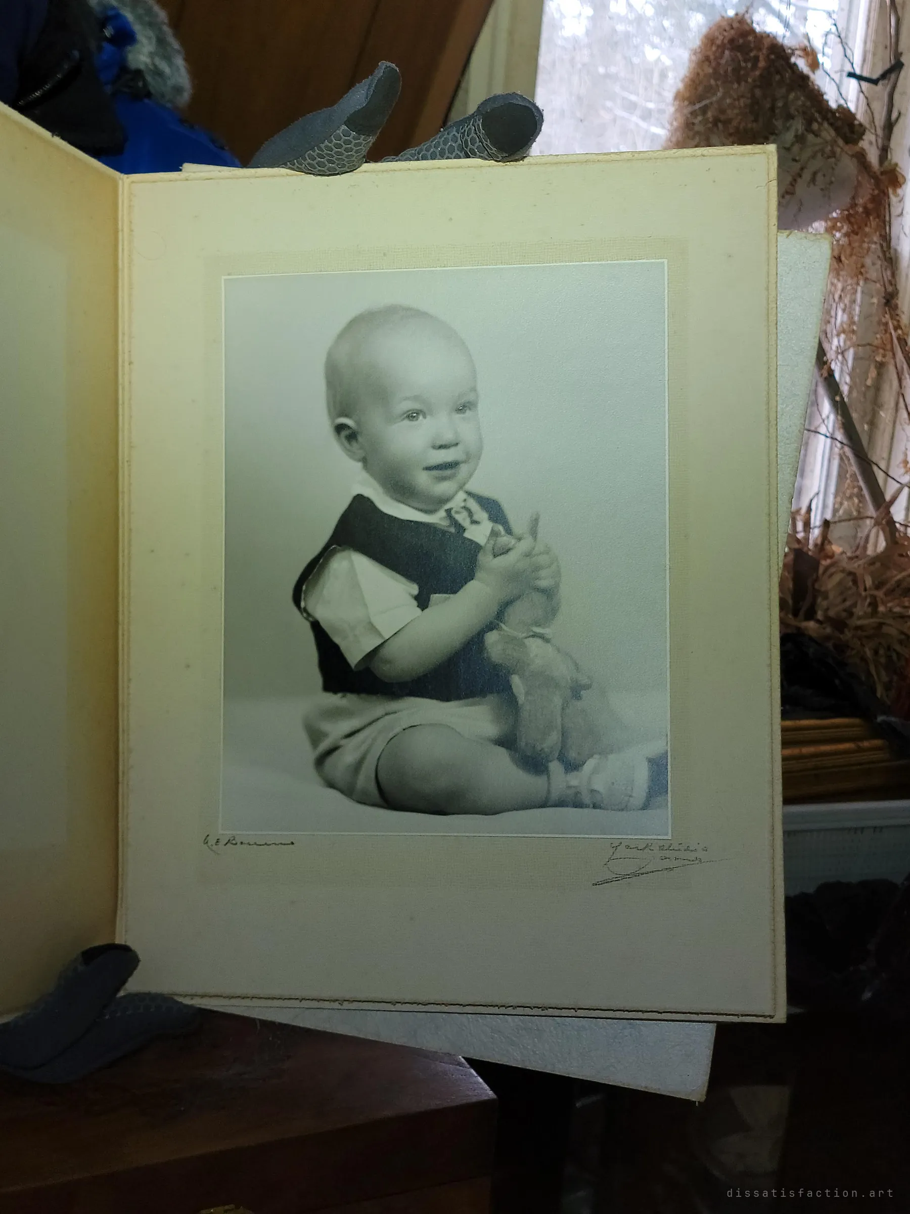 An old studio portrait of a smiling baby holding a toy, in a folding card mount beside a window with dried plants. Likely the owner as an infant, kept for decades and left behind.