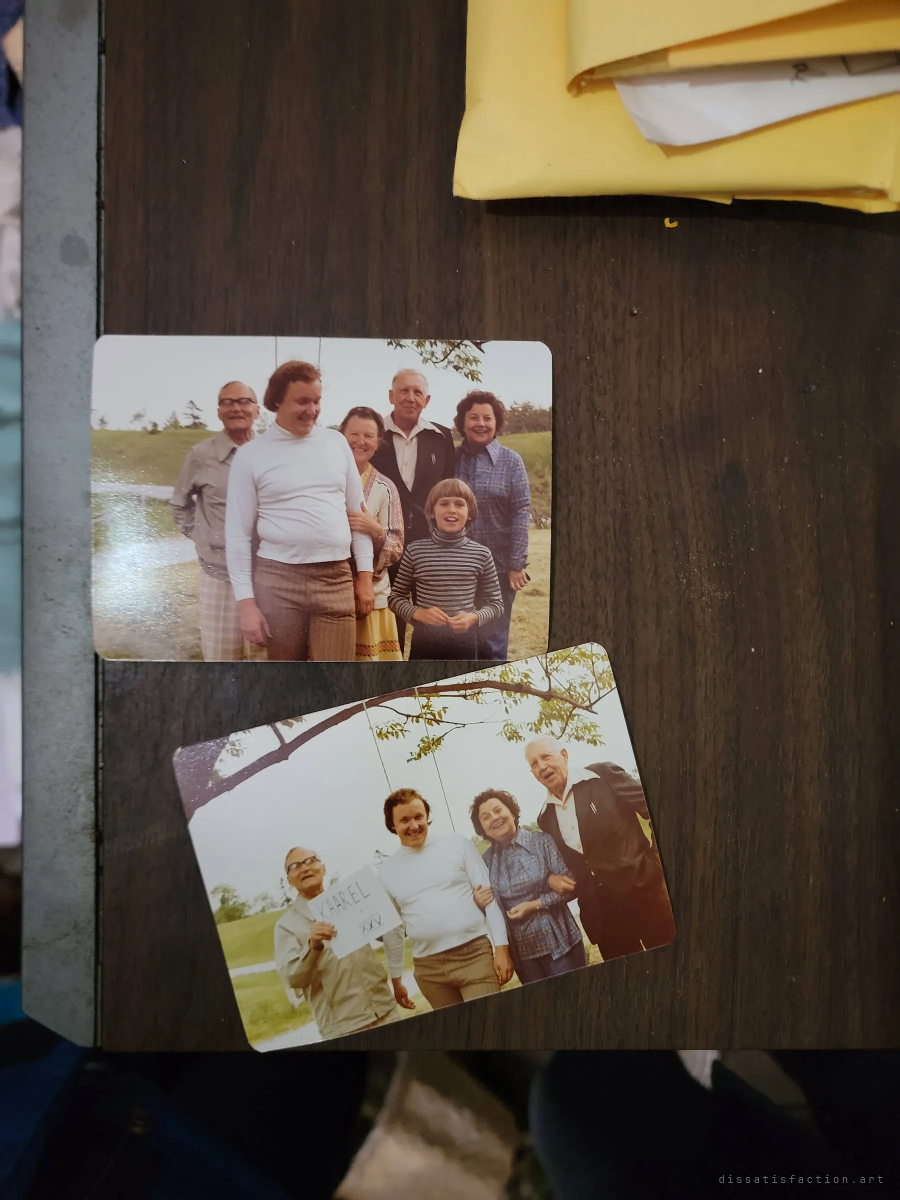 Two faded 1970s snapshots of a family posing outdoors under a tree, laid on a dark wooden table beside a bundle of photo envelopes.