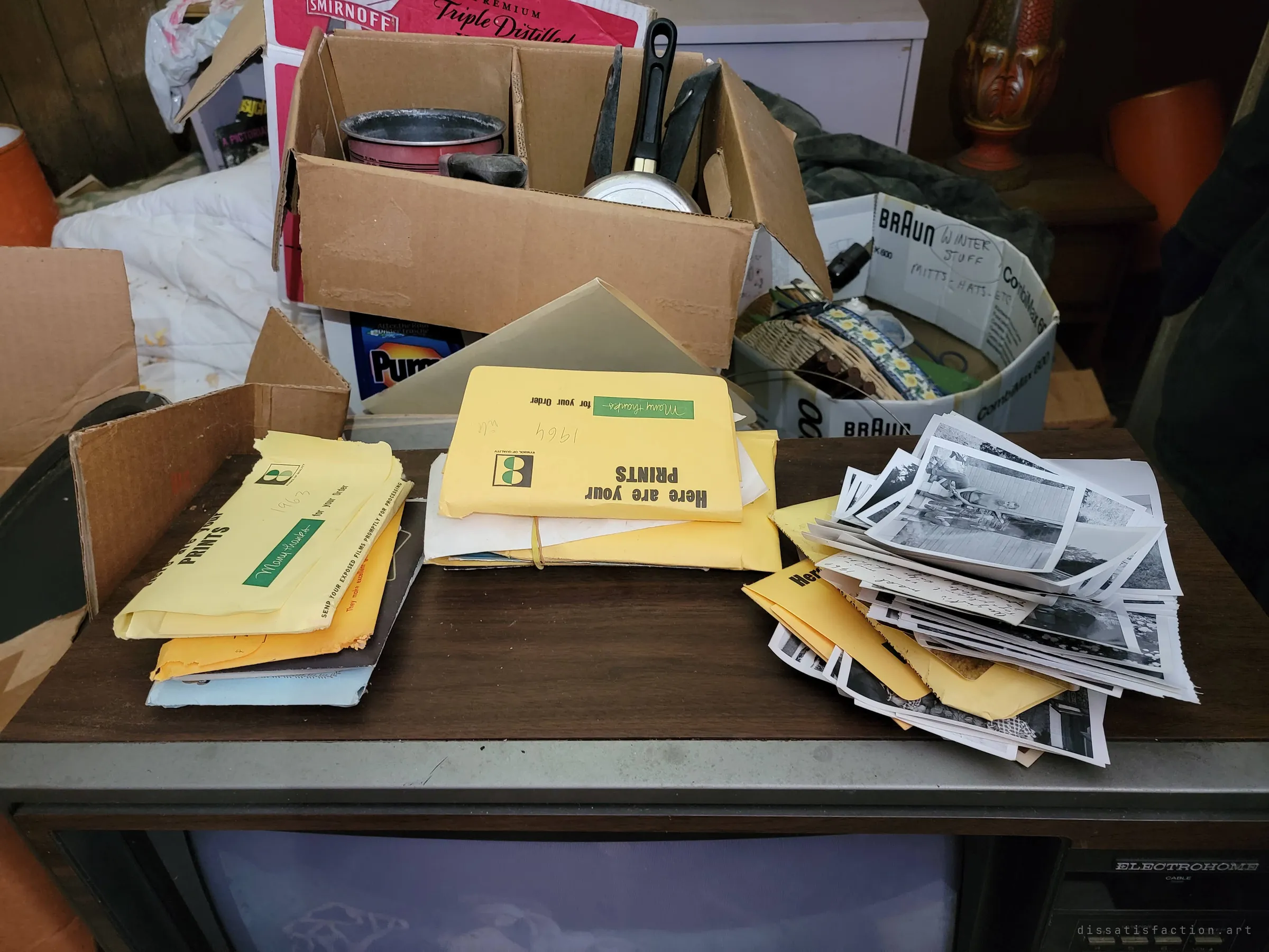 Yellow photo-lab envelopes dated from the early 1960s and a spill of black-and-white prints stacked on top of an old console television, with boxes of kitchenware behind.