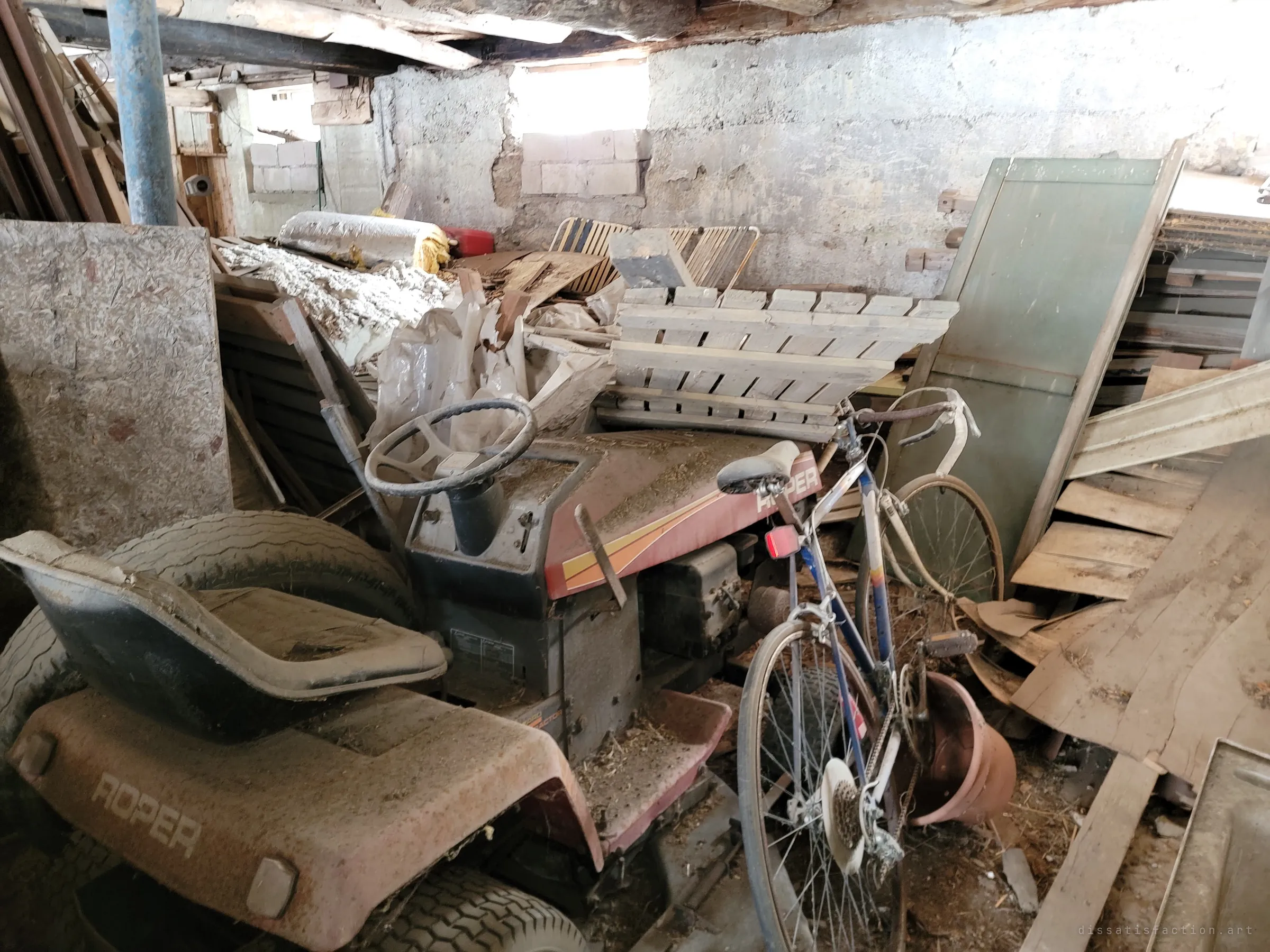 A dust-covered riding mower and an old road bike buried among boards and pallets against a crumbling stone foundation wall.