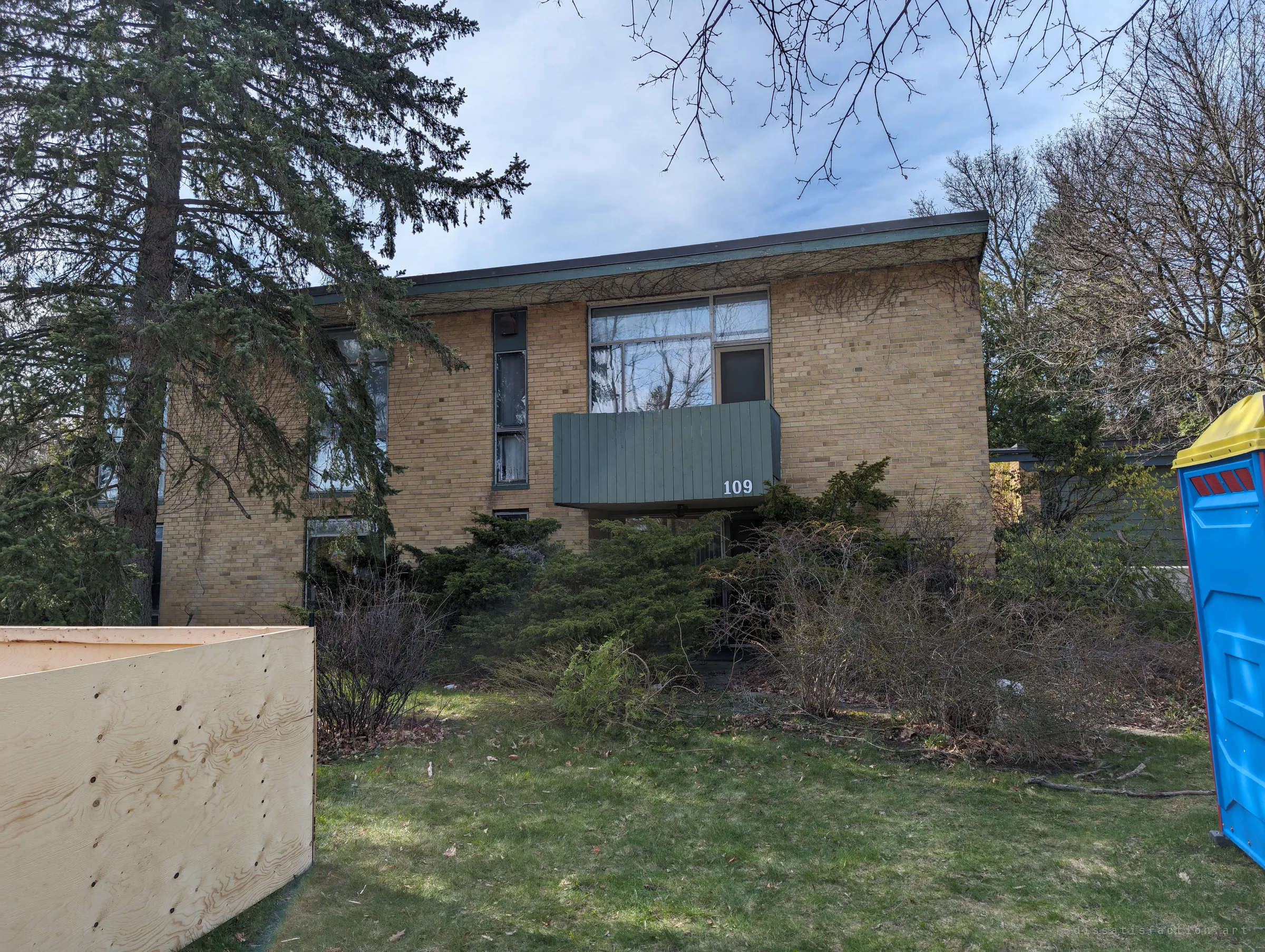 The overgrown front of a flat-roofed mid-century brick house, a green balcony over the entry, with plywood hoarding and a portable toilet on the lawn hinting at coming demolition.