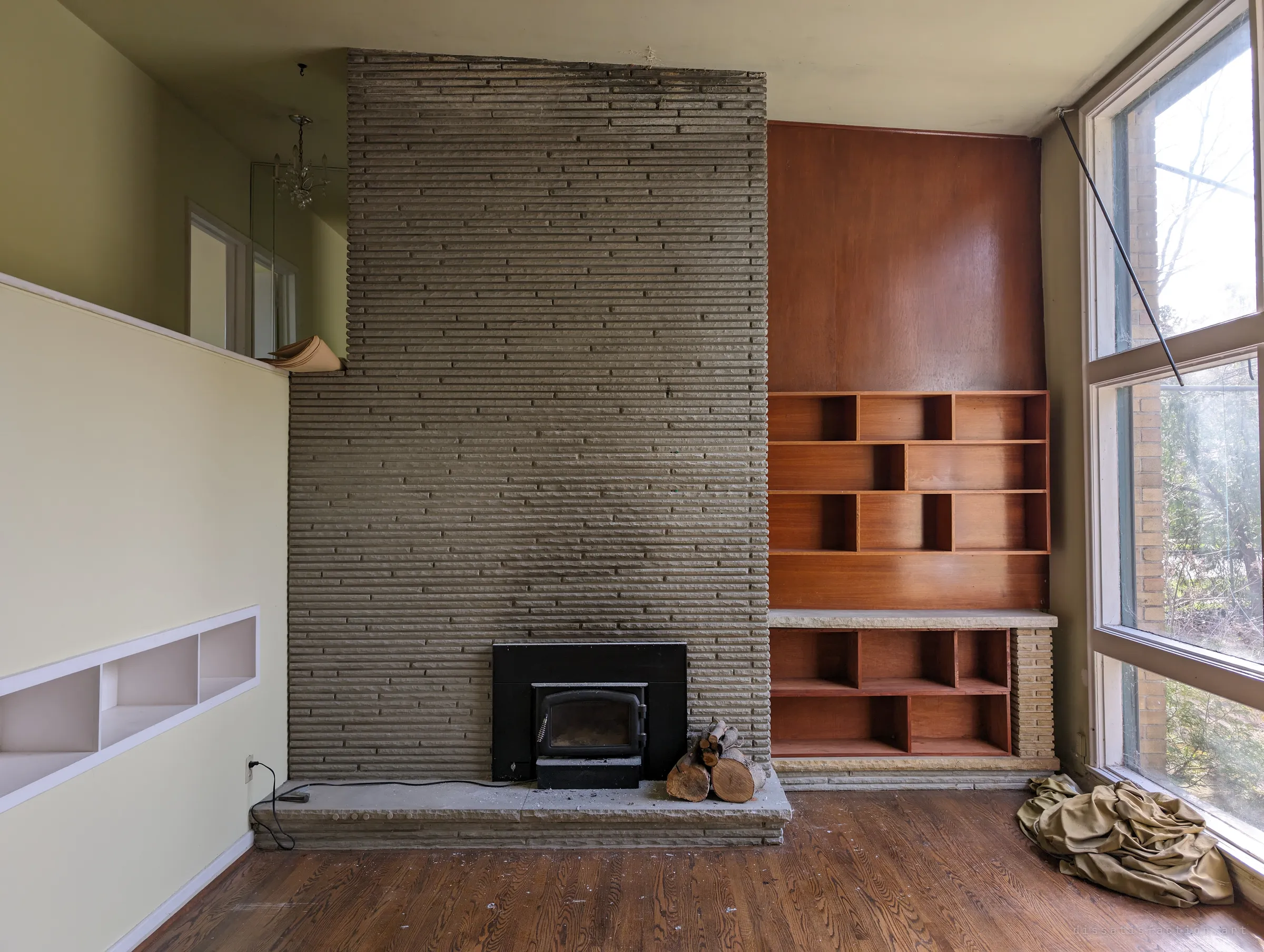A double-height living room built around a tall ribbed stone fireplace, with warm wood shelving, a stack of logs and a crumpled curtain left by the windows.