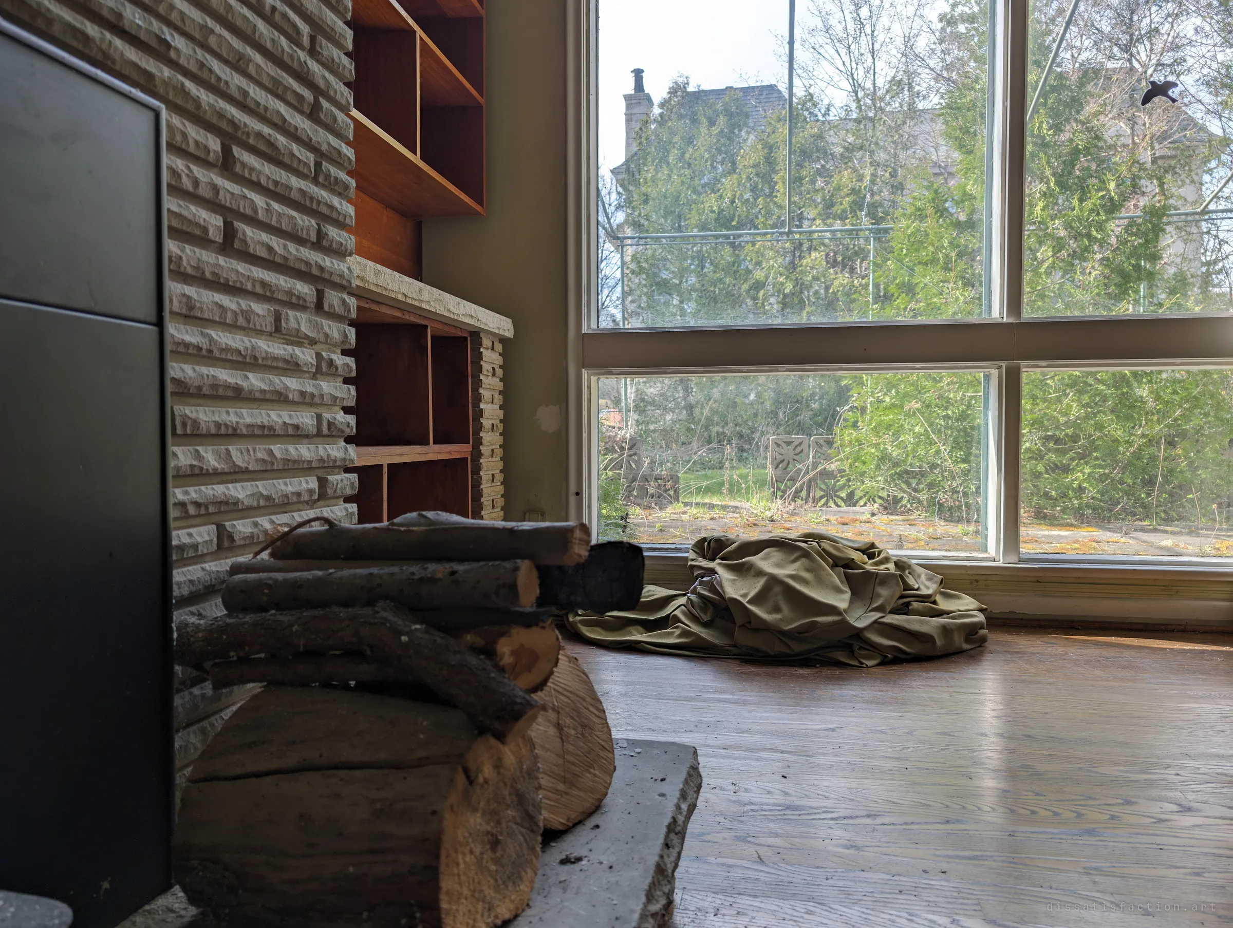 A low view past firewood and the stone hearth toward tall windows, where a discarded curtain lies in a patch of spring light.