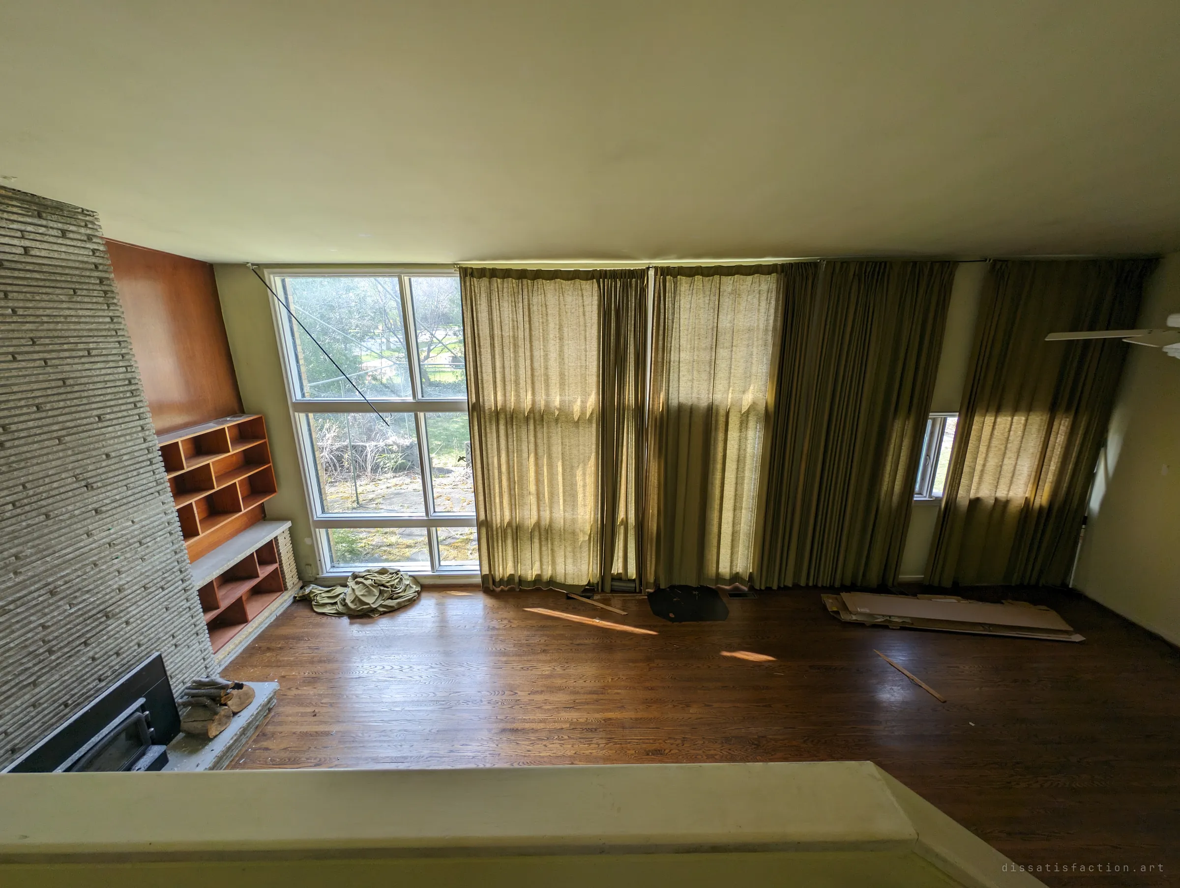 Looking down from the upper landing into the empty living room, its wall of heavy olive curtains glowing in afternoon sun beside the stone fireplace.