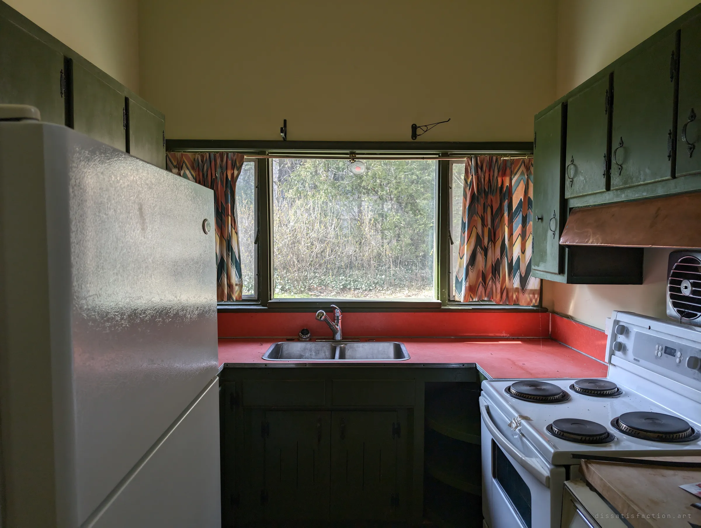 A narrow galley kitchen with dark green cabinets, a red laminate counter and chevron-print curtains framing a window onto overgrown shrubs.
