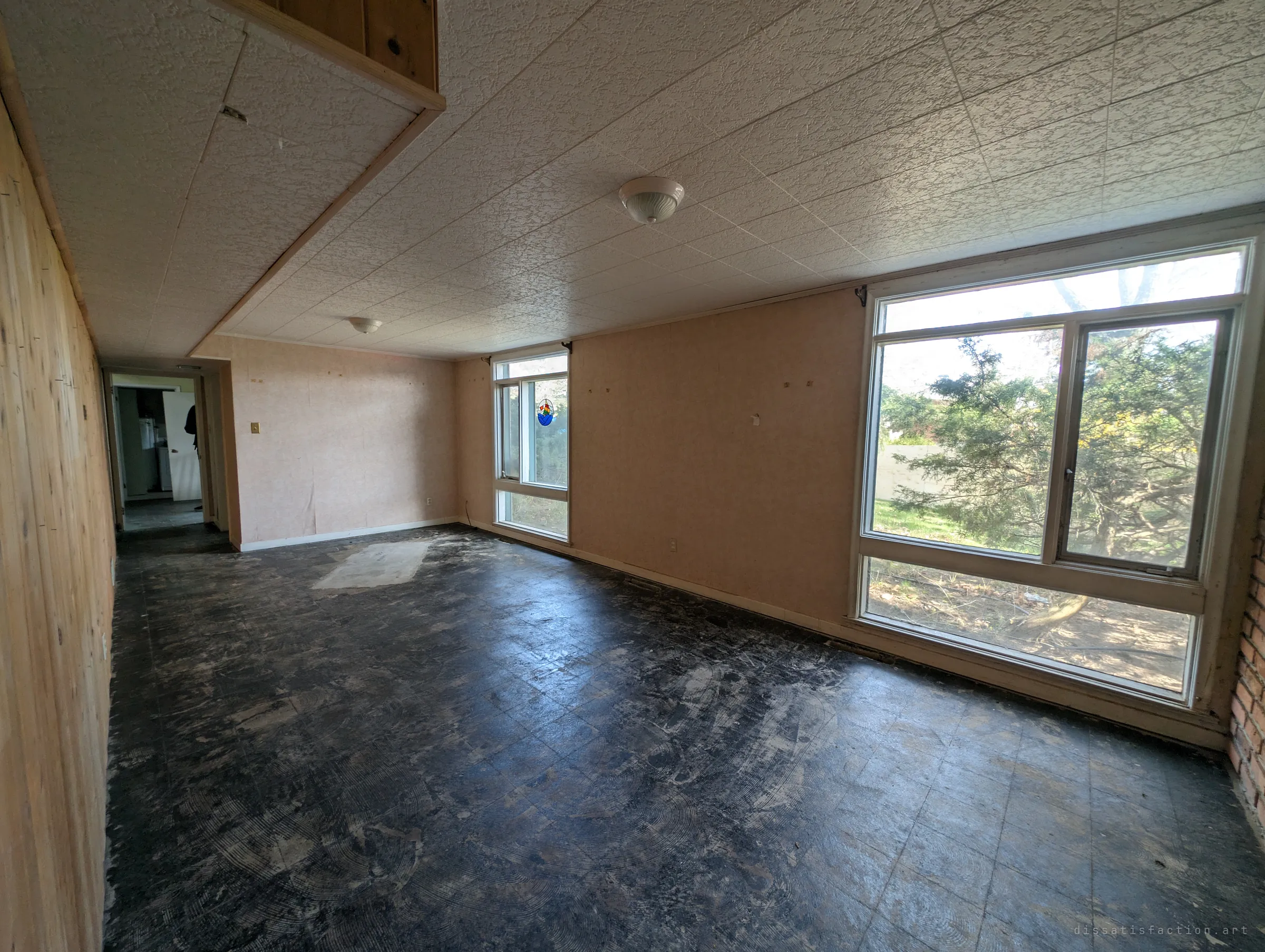 A long lower-level room stripped to its black tile adhesive, pink wallpaper and bare windows looking out on an untended yard.