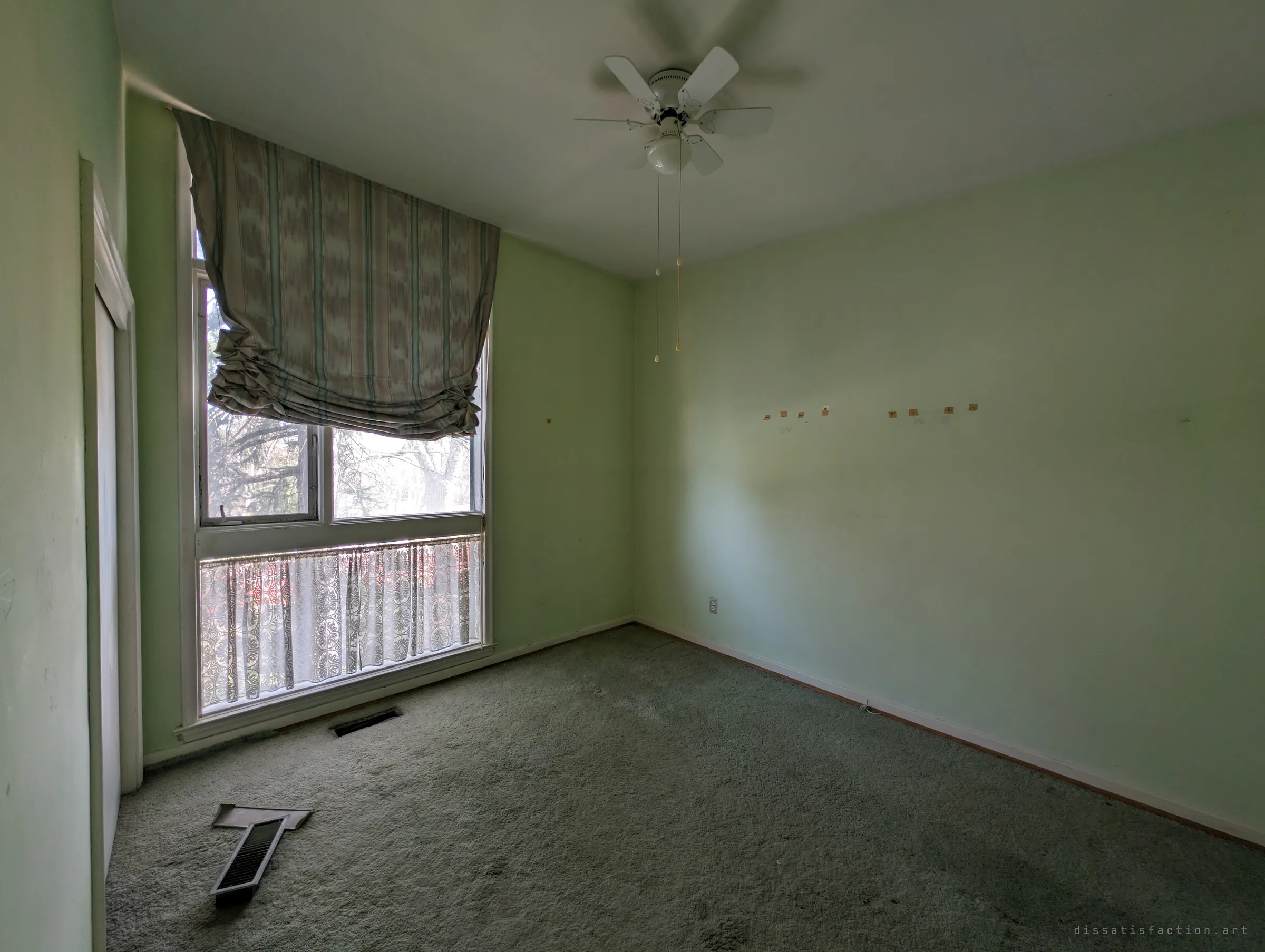 An empty mint-green bedroom with a sagging patterned blind half over a tall window and a ceiling fan still hanging. Pulled-up floor vents and tape scars on the walls show the room was stripped and left.