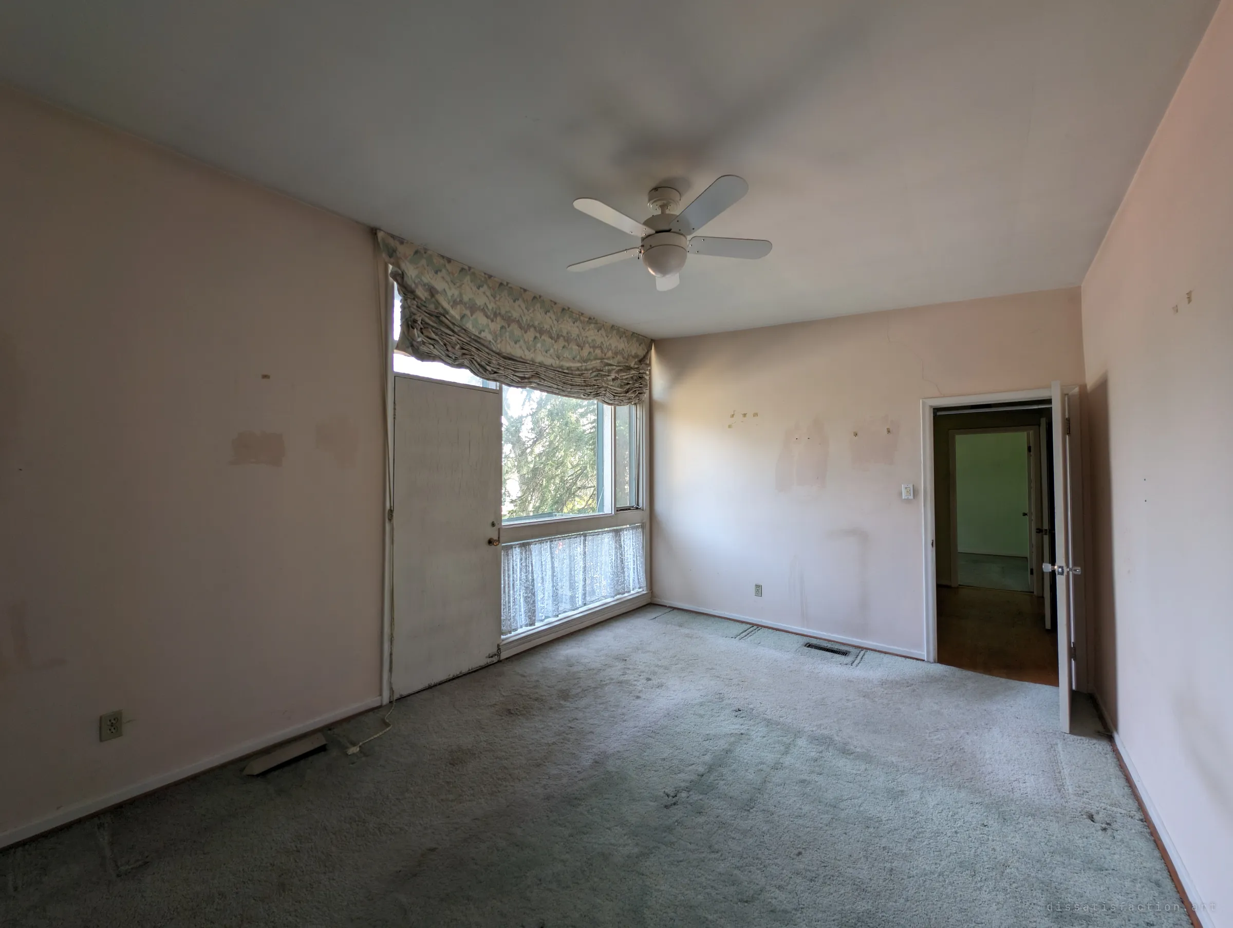 A pale pink bedroom with worn carpet, a ruched blind over a corner window and a boarded panel beside it. Through the open door a hallway leads to another green room, hinting at the house's layout.