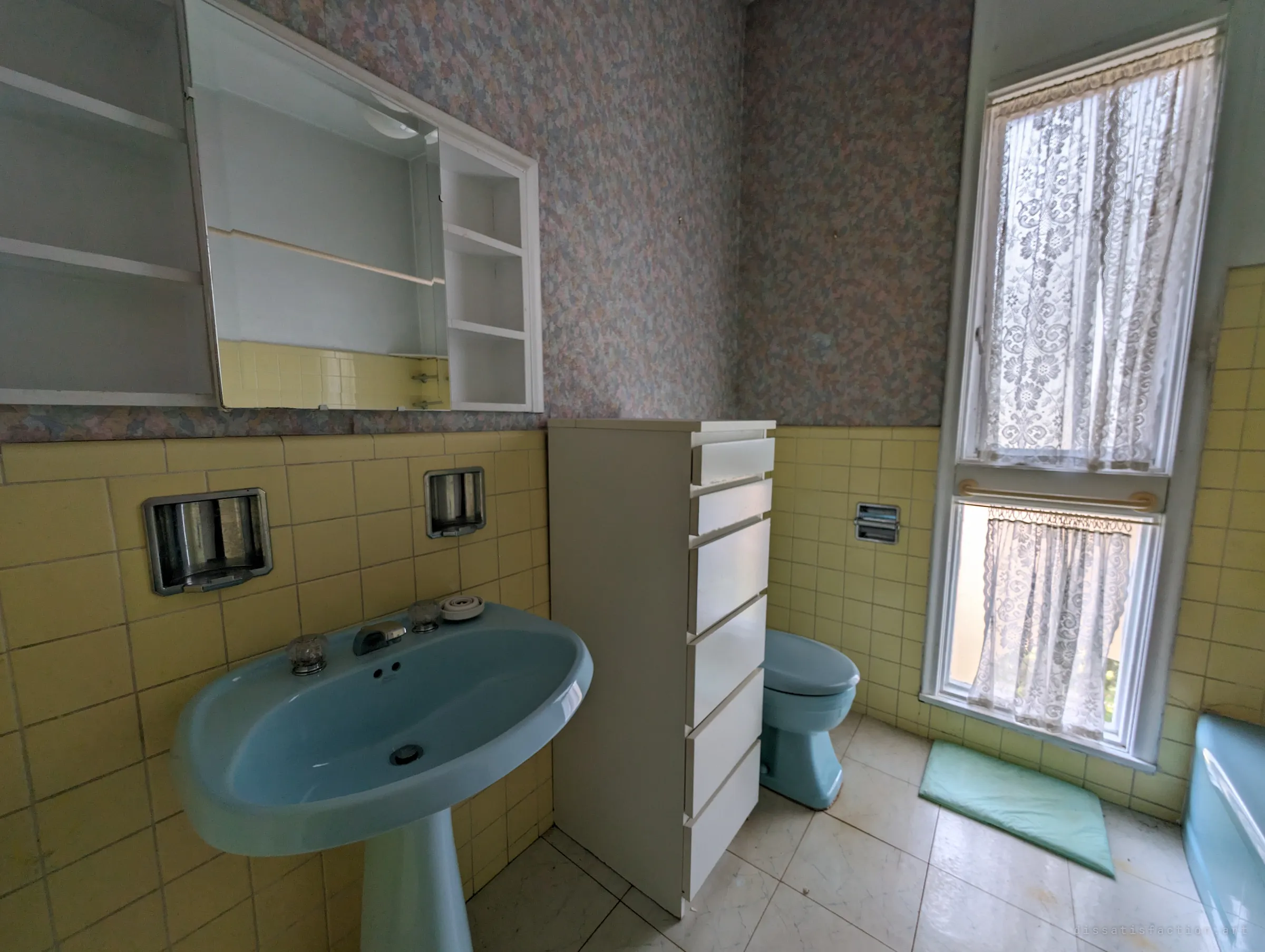A mid-century bathroom with a powder-blue pedestal sink, toilet and tub against butter-yellow tile and speckled wallpaper. Lace curtains still hang in the tall window, a time capsule left intact.