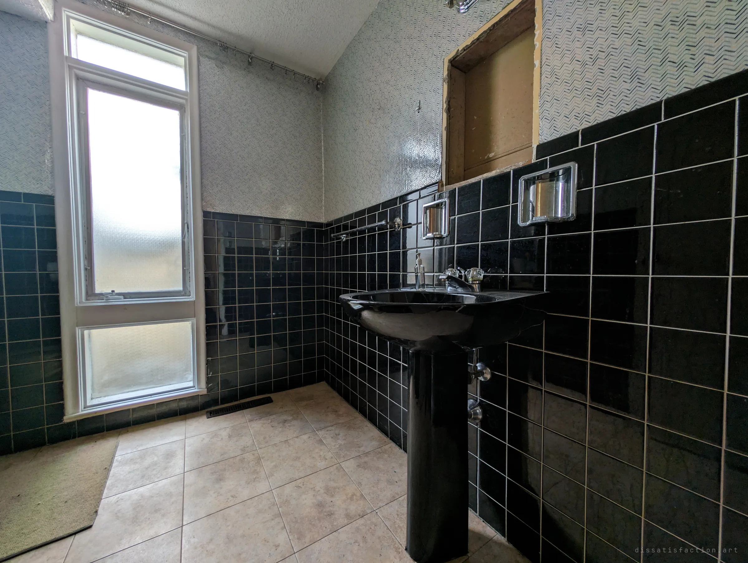 A second bathroom done in glossy black tile with a black pedestal sink and chevron wallpaper, light coming through a frosted window. An empty cabinet recess above the sink shows where fittings were pulled out.